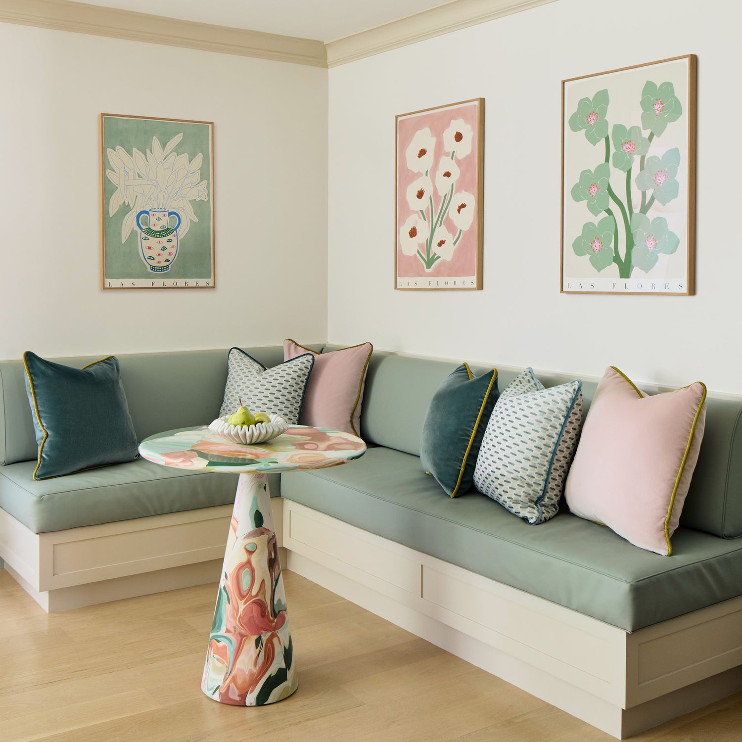 blue green cushion at a kitchen bench with rose pink velvet pillows, peacock blue velvet pillows, and mint green geometric pillows on the bench with a multicolored table in front of the bench and floral pictures hung on the wall above the bench