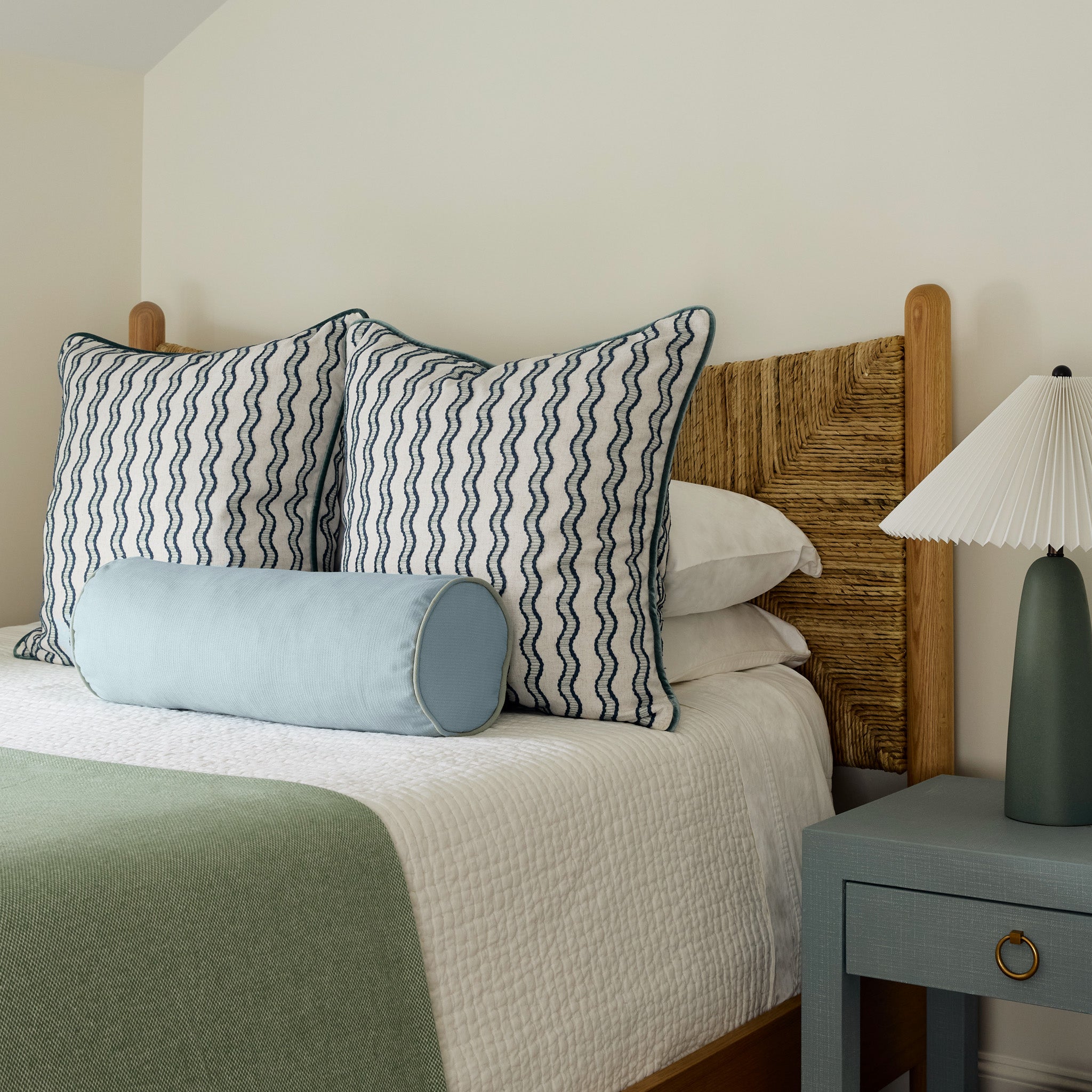 embroidered cream and navy blue wavy lines and grey blue pillows on a bed with white and sage green bedding and a light blue grey night stand with a green and white lamp on it