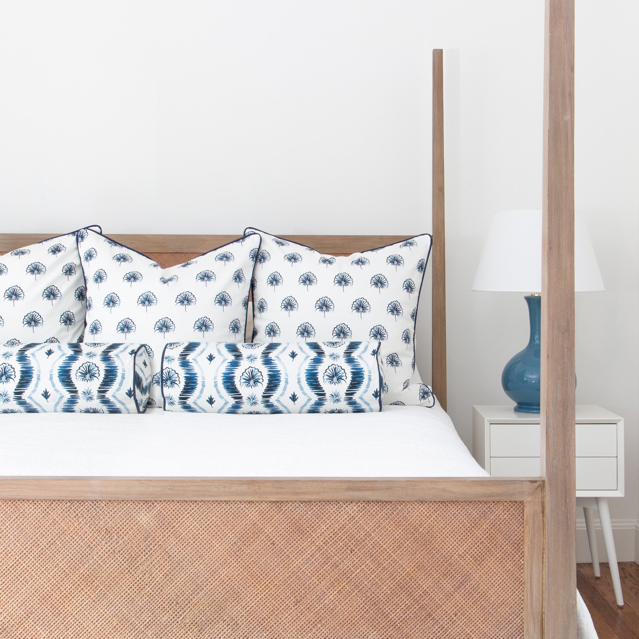 White bed with three Blue Ikat customed pillows and two customed Blue Ikat Striped bolster pillows by white nightstand with a blue lamp on top