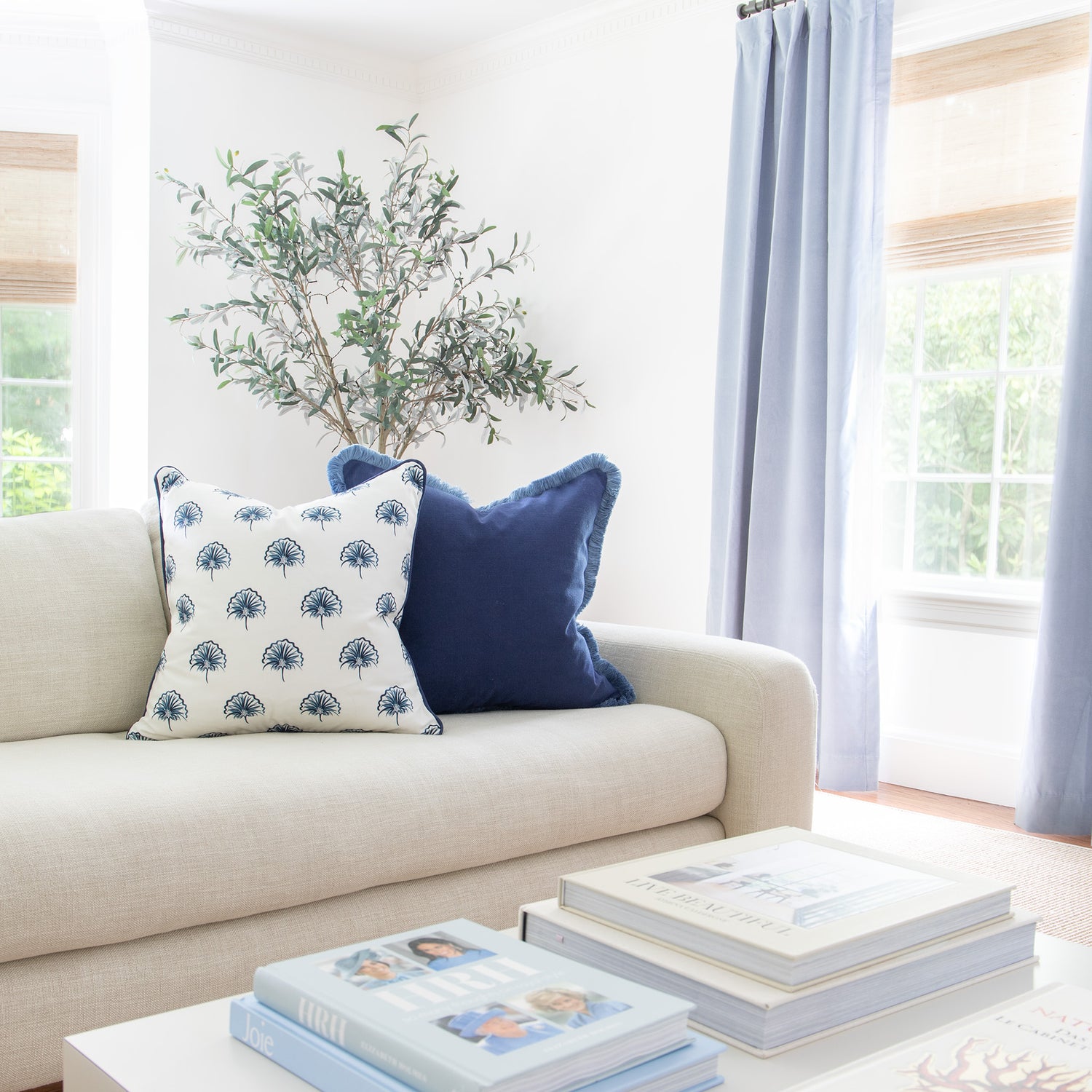 Living room styled with Sky Blue Velvet Curtains by illuminated window next to beige couch with Floral Navy Printed Pillow with Navy Piping and Navy Blue Pillow with Navy Fringe
