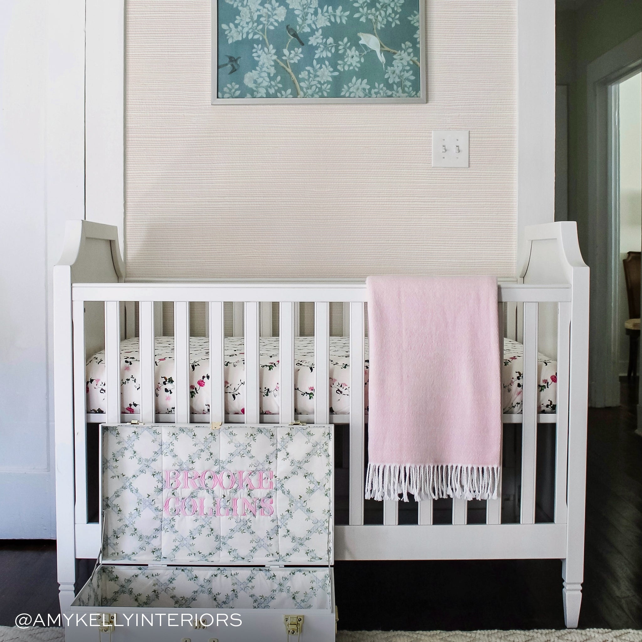 close up photo of a nursery with a white crib with a pink blanket on it. pale pink grasscloth wallpaper on the wall and a blue botanical framed photo on the wall above the crib 