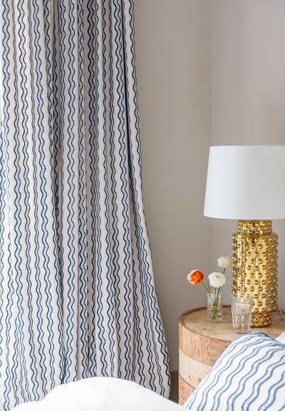 cream curtains with wavy blue line pattern on them with a wooden night table next to the curtains with a gold lamp on it and a vase of flowers 