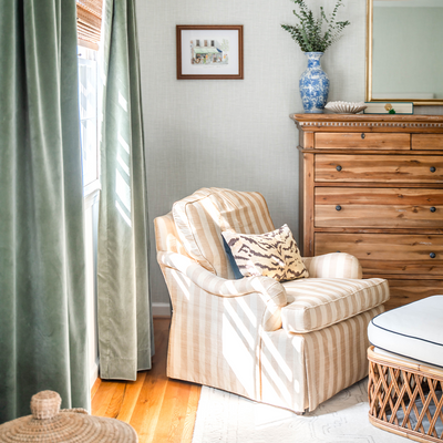 blue and green velvet curtains hung in front of an illuminated window with a beige striped chair with an animal print pillow on it and a brown wooden dresser