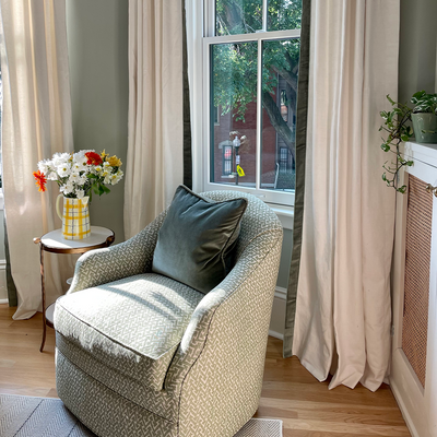 white curtains with fern green velvet band trim in front of a window with a green chair in front of the window with a green velvet pillow on the chair and a white side table next to the chair with a yellow vase with flowers in it