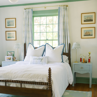 Blue & Green Striped curtains in front of an illuminated window in a bedroom with a bed in front of the window with white pillows and Blue & Green Striped pillow on the bed and a blue night stand next to the bed