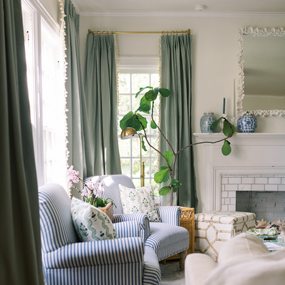 sage green curtains hung in front of an illuminated window with a white tiled fireplace next to the window and a blue and white striped chair 