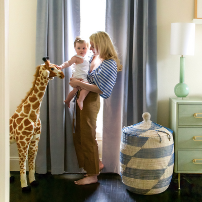 sky blue velvet curtains hung in front of an illuminated window with a mom holding her baby in front of the window and a large stuffed animal giraffe and a white and blue basket