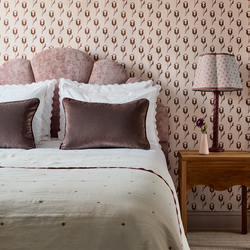 neutral background wallpaper with pink and maroon abstract floral pattern on it in a bedroom with a pink velvet headboard on the bed with white bedding and white pillows with brown velvet pillows and a brown wooden nightstand with a lamp on it  