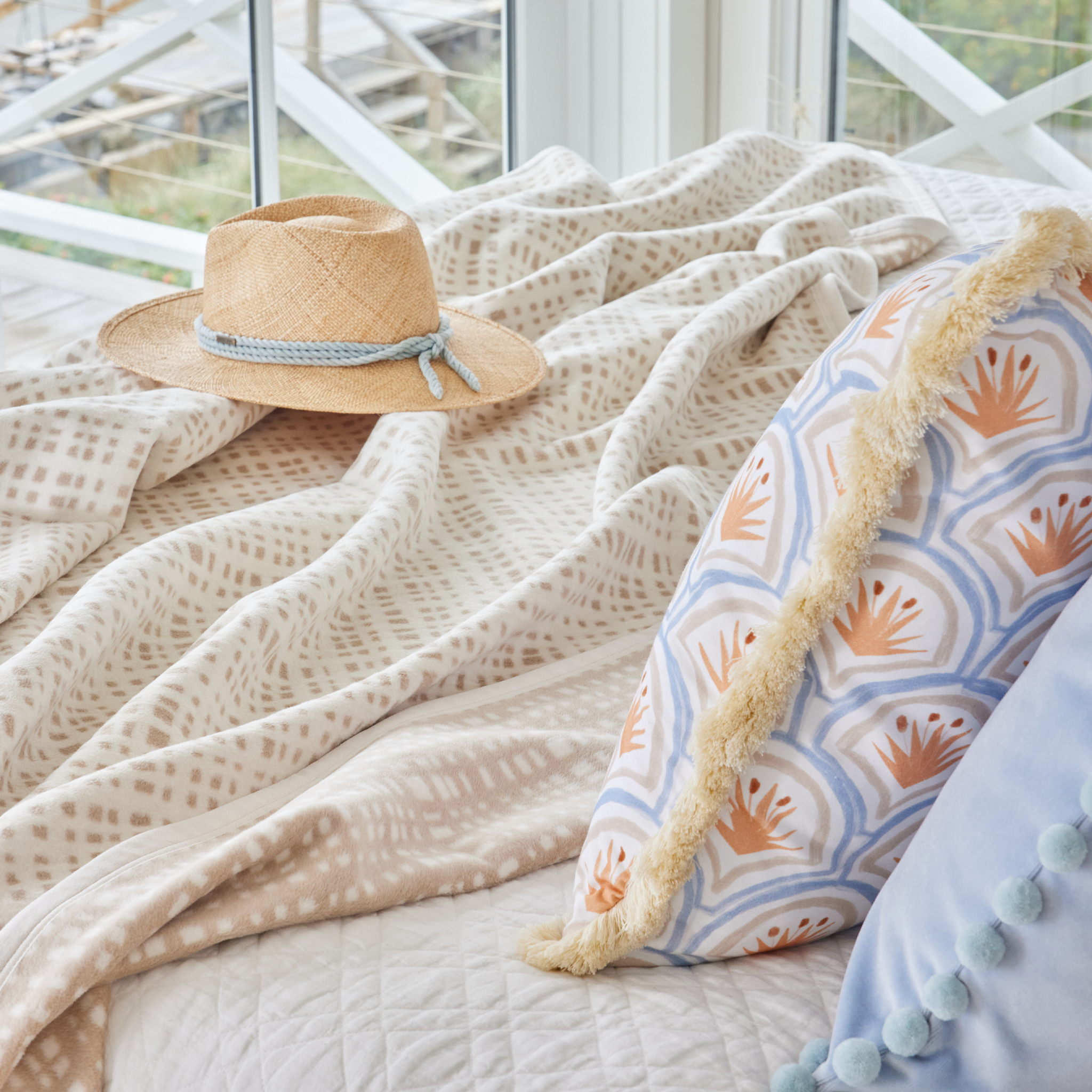 Neutral bed close-up styled with Art Deco Palm Pattern Printed Pillow and Sky Blue Velvet Pillow with Pom Poms next to a gingham blanket and straw hat.