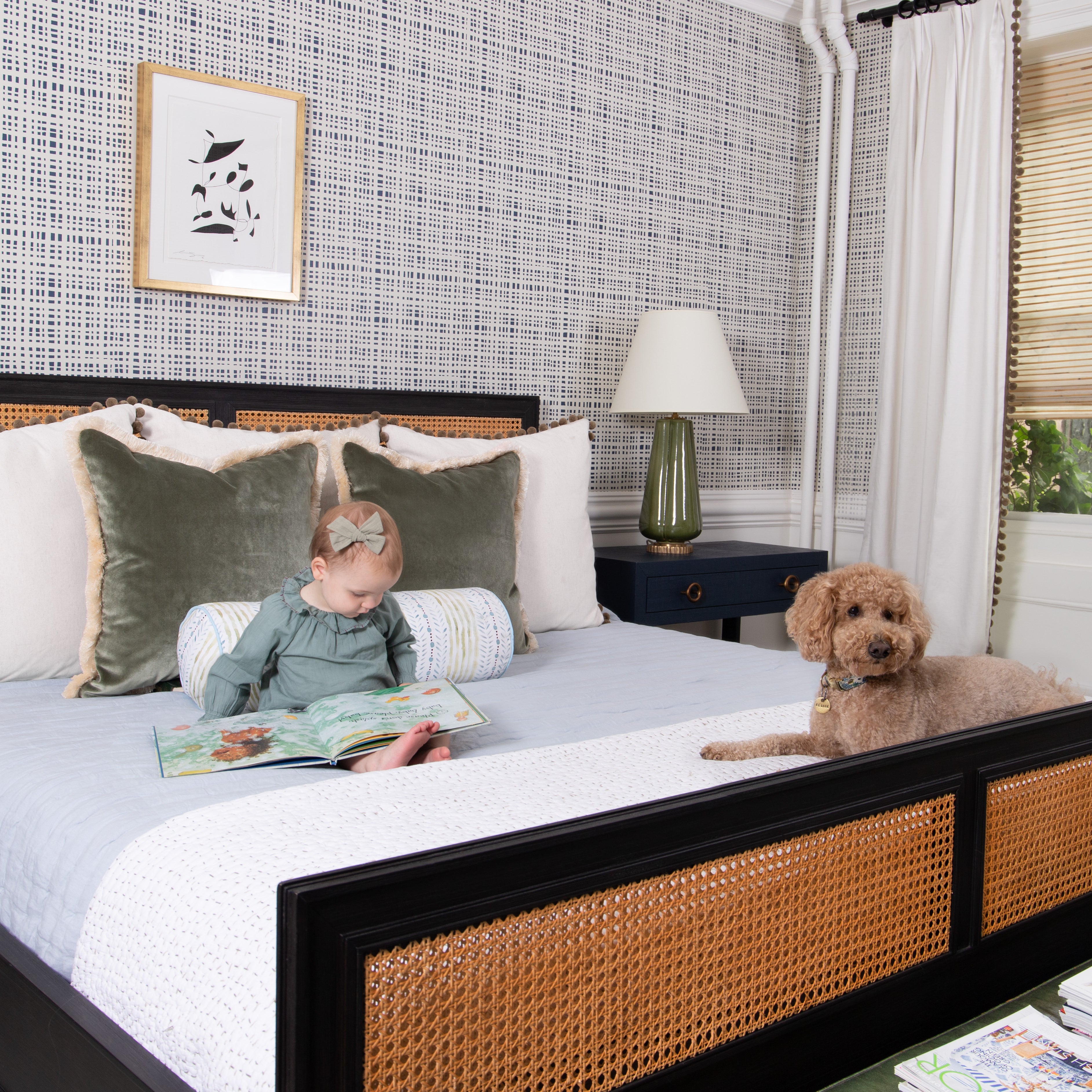 Bedroom styled with blue nightstand white and green lamp on top next to Natural White Linen Curtains with brown pom poms with two Fern Green Velvet Pillows on white bed by baby in green dress reading next to brown dog with Navy Gingham Printed Wallpaper with a black and white artwork hung