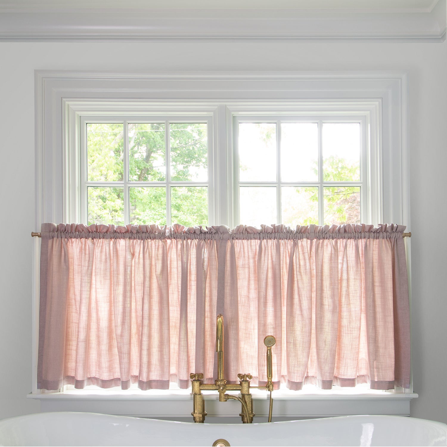 light pink cafe curtain hung in front of an illuminated window in a bathroom with a white bathtub in front of the window with a gold faucet 