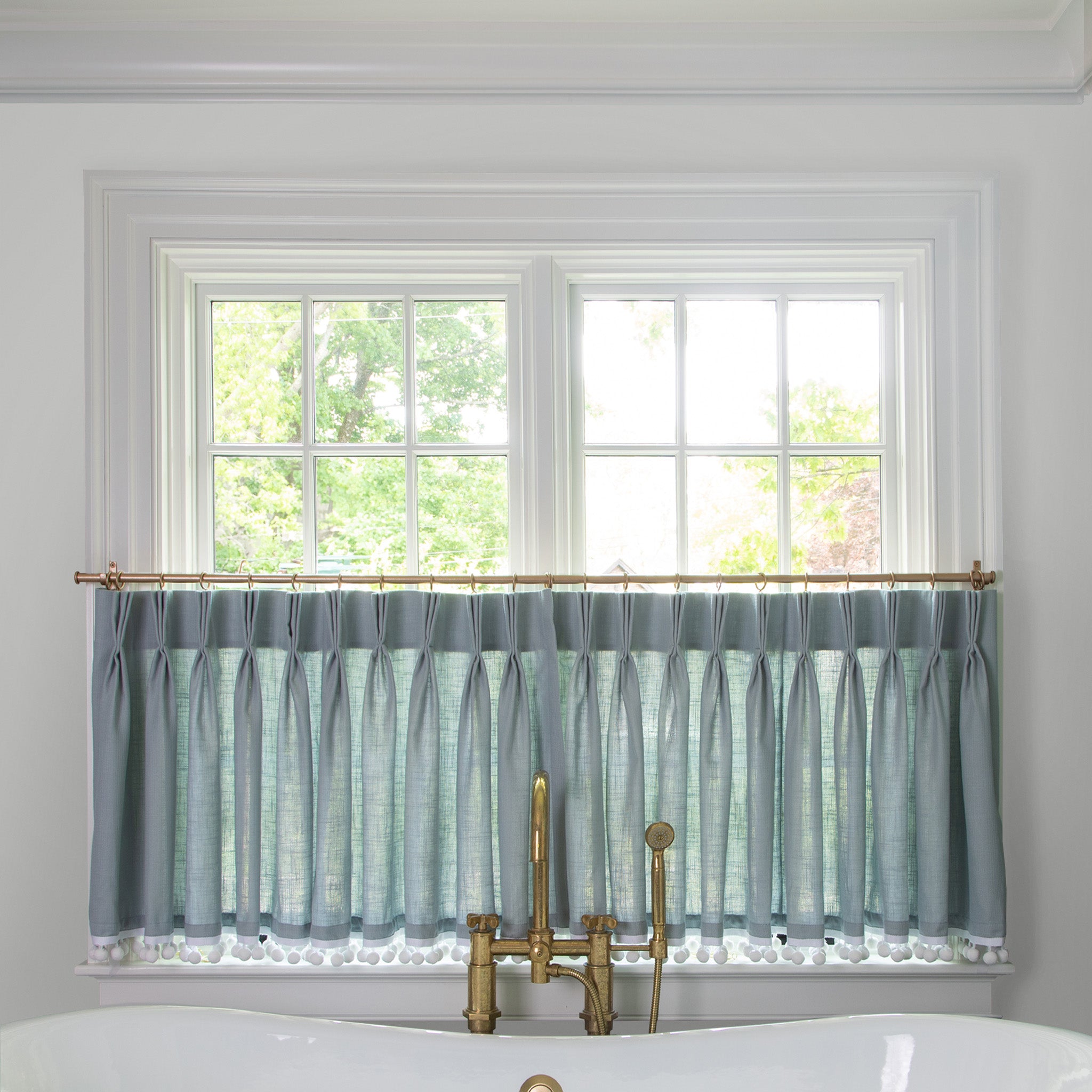 grey blue  cafe curtain hung in front of an illuminated window in a bathroom with a white bathtub in front of the window with a gold faucet 