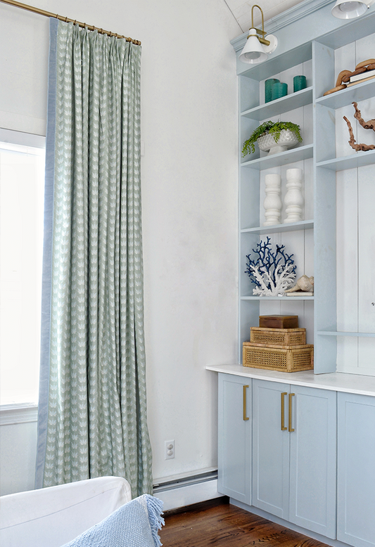 Blue & Green Floral Drop Repeat curtains hung in front of an illuminated window in the corner of a room with light blue cabinet and shelves with rattan boxes and blue and white coral and candles on the shelves