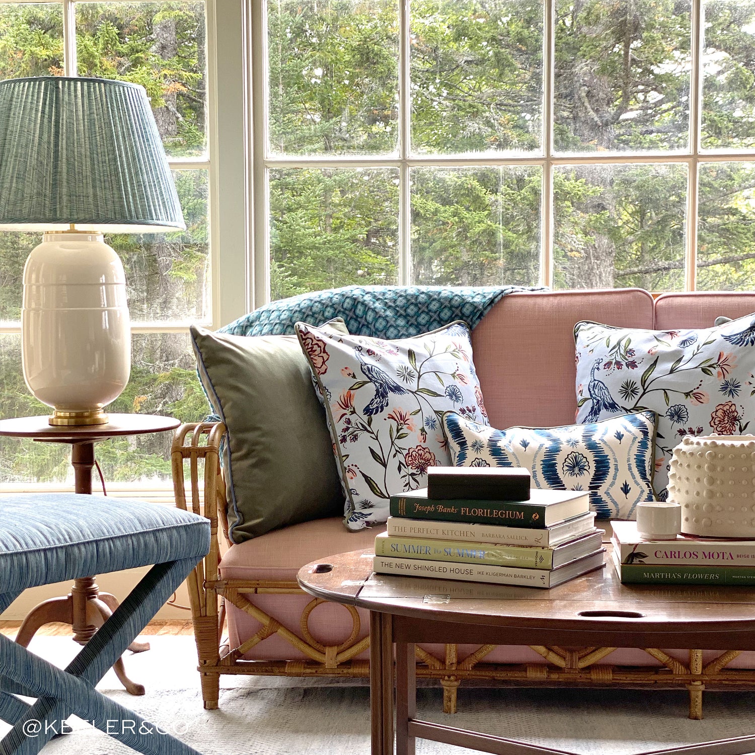 In front of a clear window with a Coral Couch with Fern green pillow, daphne powder pillow and blue itak pillow besides a wooden small table and green and white lamp on top. Wooden circular coffee table in front of couch with two stacks of books. Photo taken bye Keeler & Co.