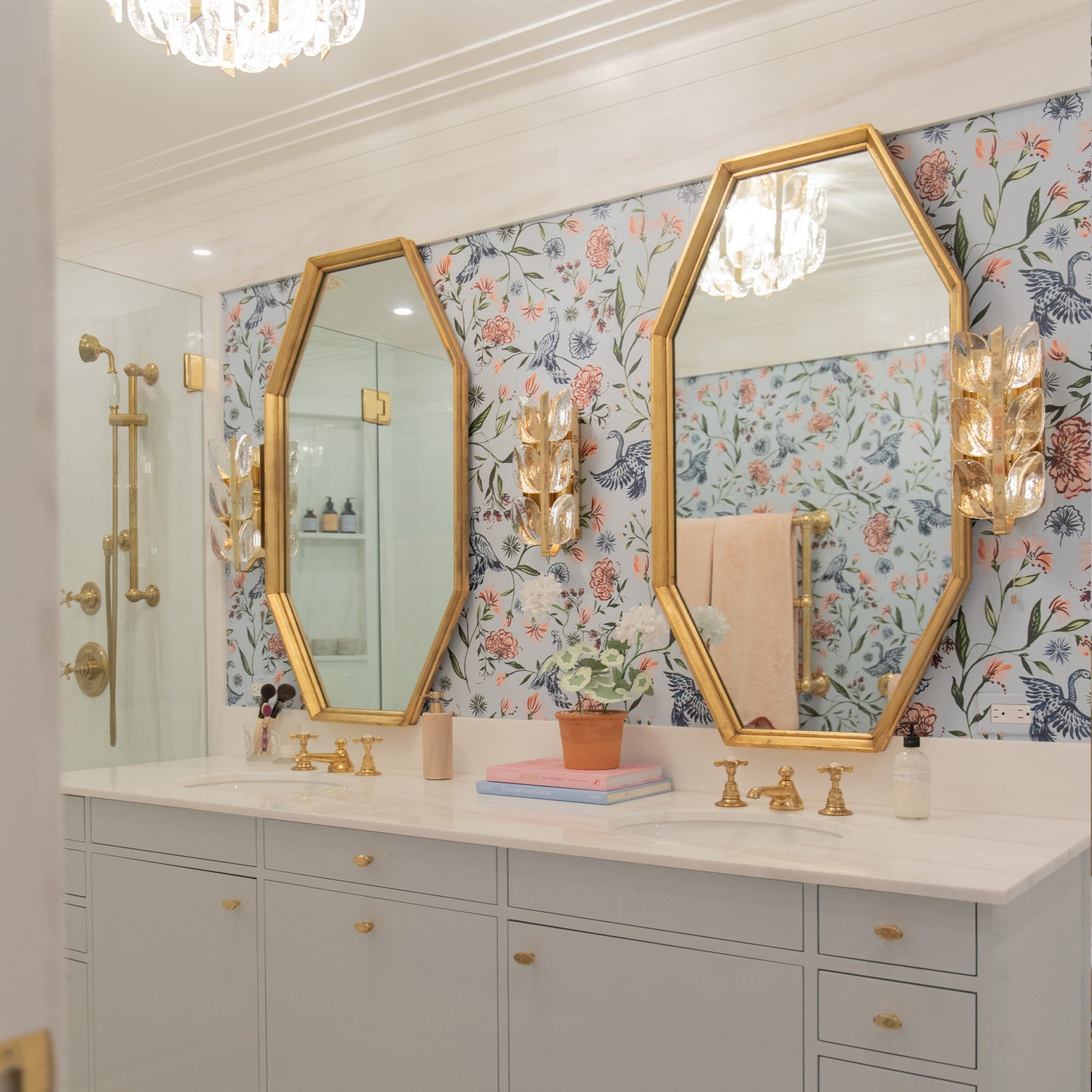 Illuminated bathroom styled with Blue Chinoiserie Printed wallpaper with two gold framed mirrors on top of two white sinks.