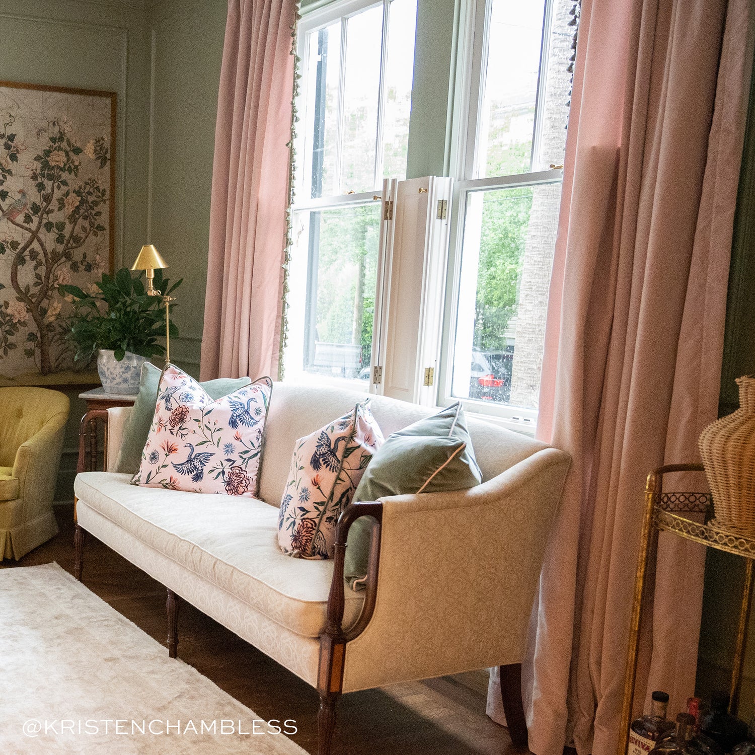 Living room styled with two Pink Chinoiserie Pillows facing each other and two fern green pillows by them on cream couch with window to the back styled with pink curtains with green pom pom fringe. Photo taken by Kristen Chambless
