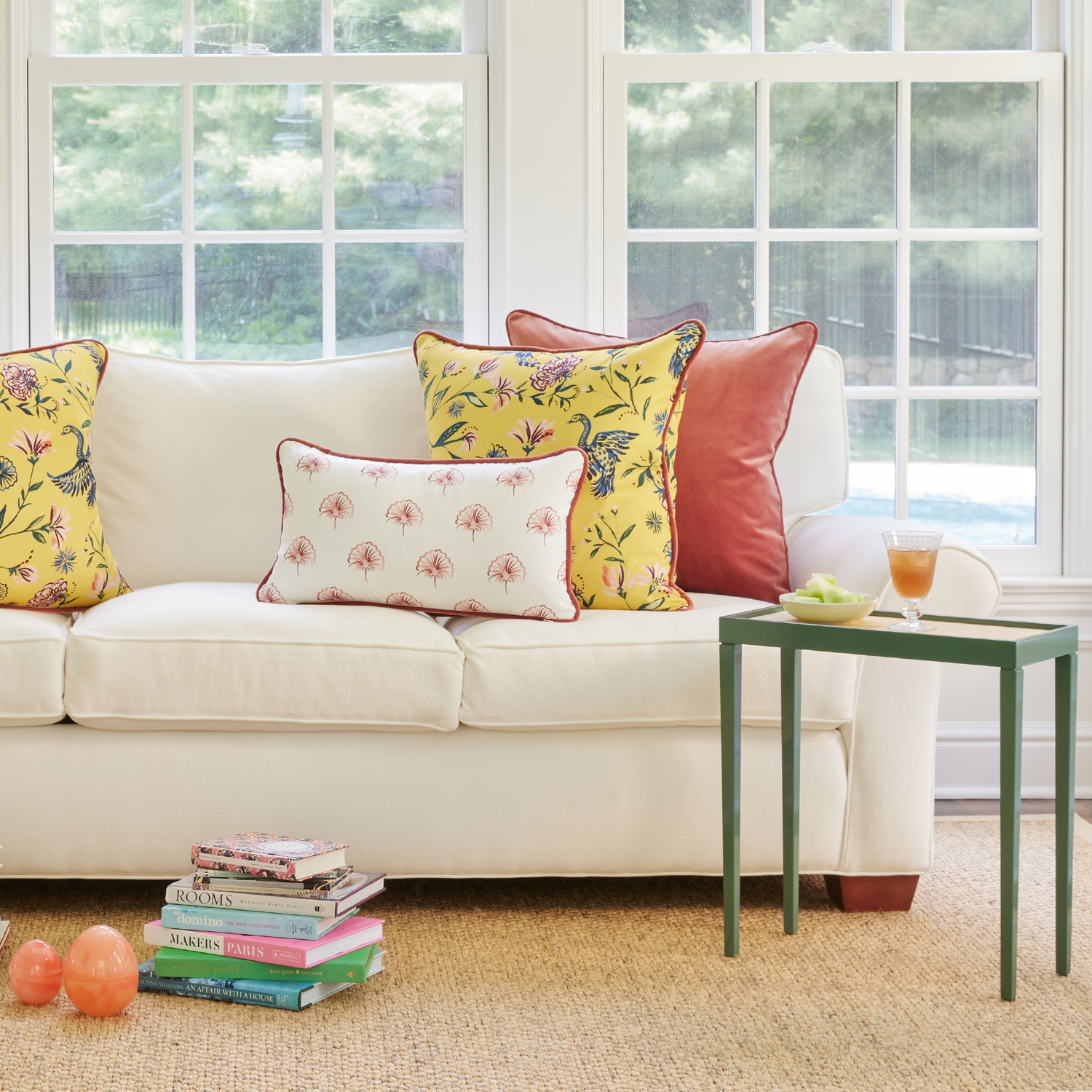 White Couch with Floral Pink, Yellow Chinoiserie, and Coral Velvet pillows with Green table with plate on top with two stacks of books on the floor