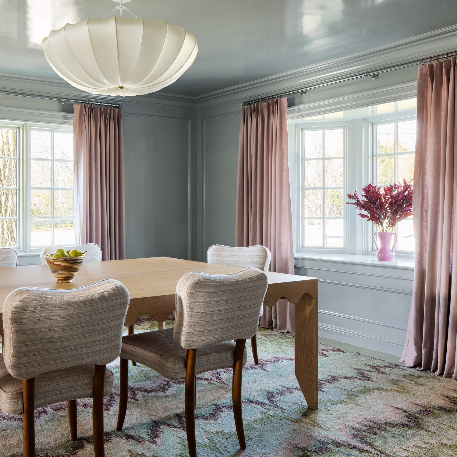 soft pink curtains on a metal rod in front of an illuminated window in a dining room with a wooden table and white chairs