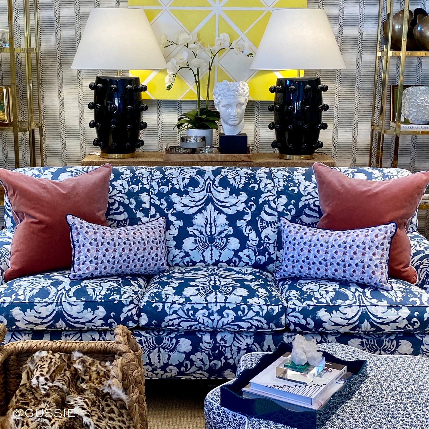 Living room styled with coral velvet pillows and two red and blue printed lumbars on blue printed couch under two black and white lamps with home decorations. Photo taken by Gussie
