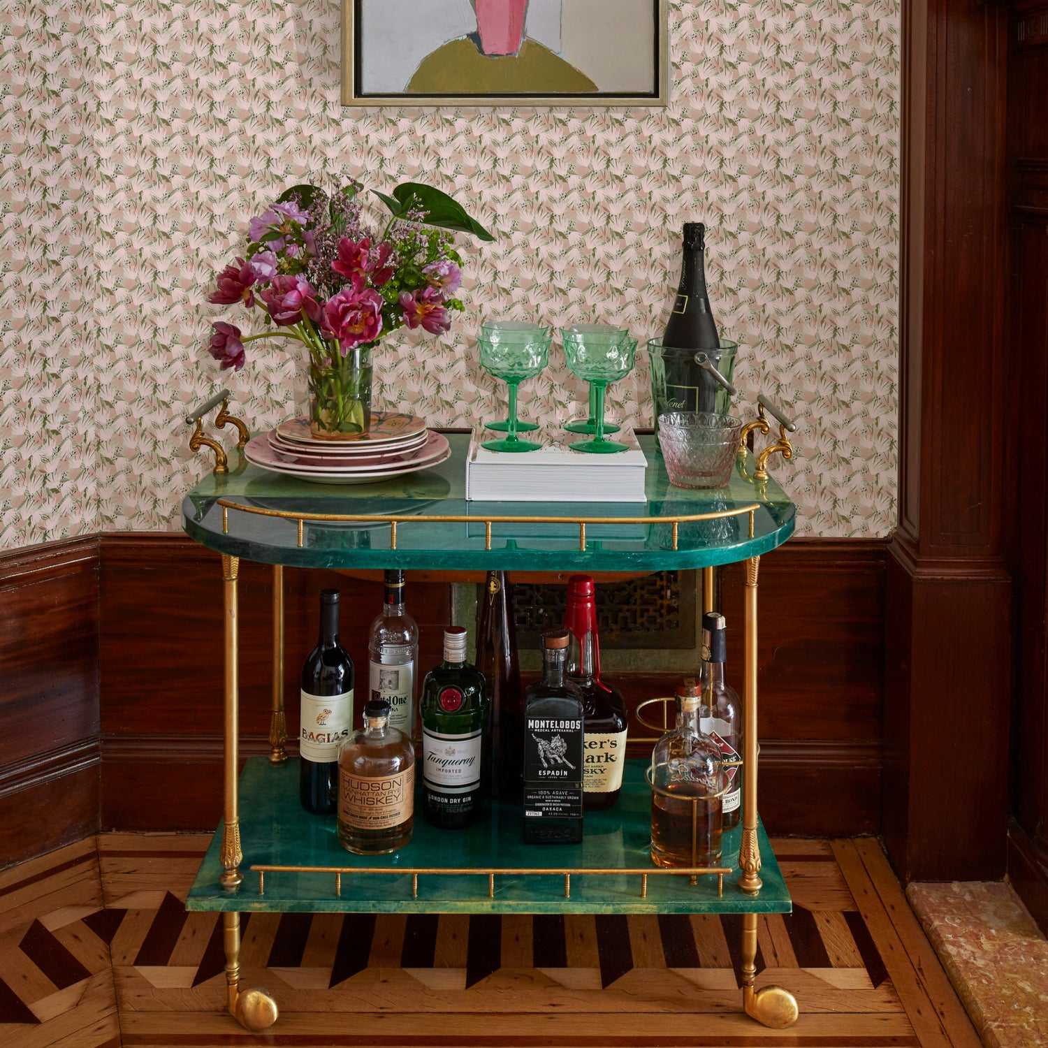 Green and gold bar cart with liquor in the bottom shelf and green glasses on top of a white book by pink flowers on stacked pink plates in front of Pink Floral Printed Wallpaper