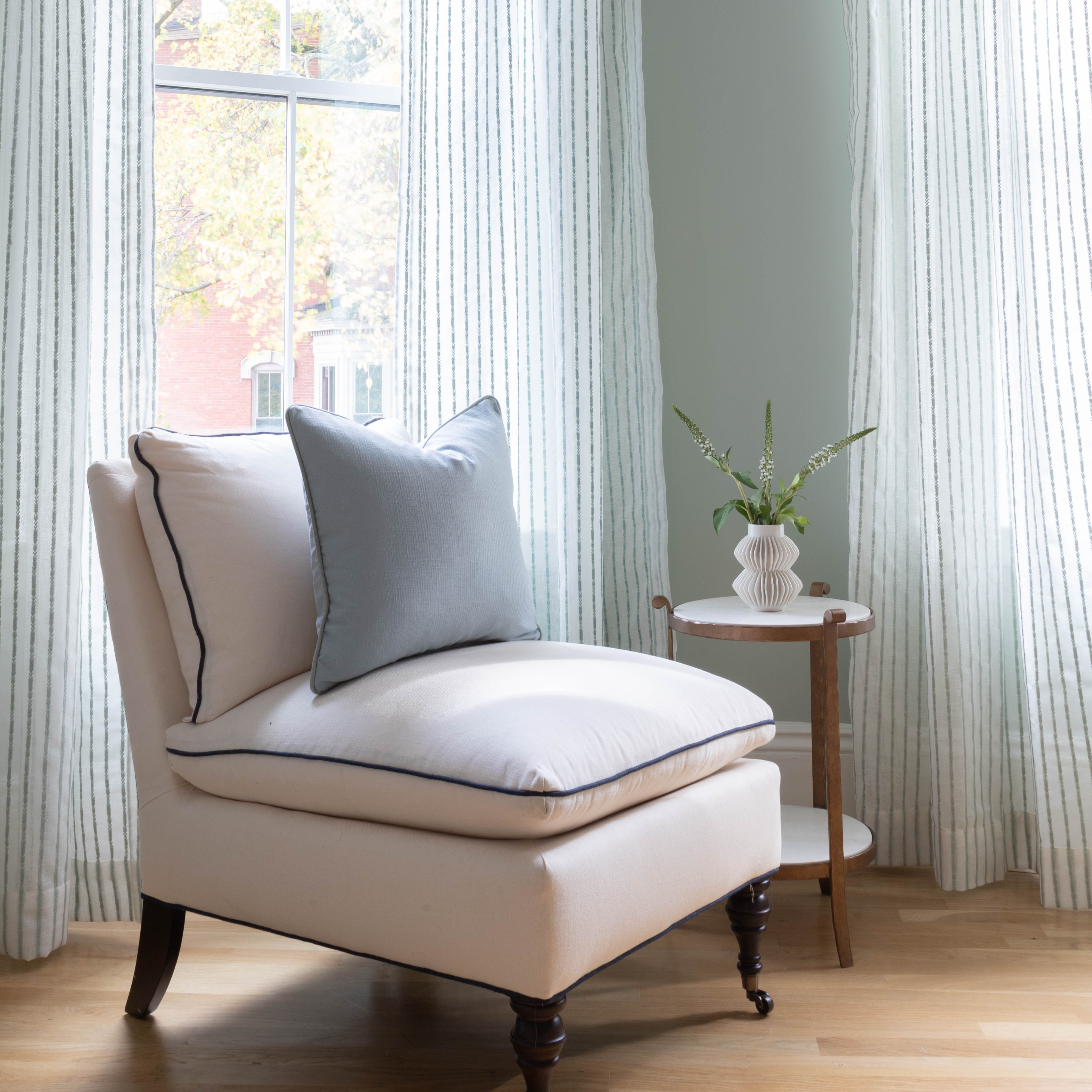 sheer white curtains with mint green embroidered stripes on them hung in front of an illuminated window with a white chair with a blue pillow on the chair sitting in front of the window with a brown and white side table next to the chair with a white vase with plants in the vase