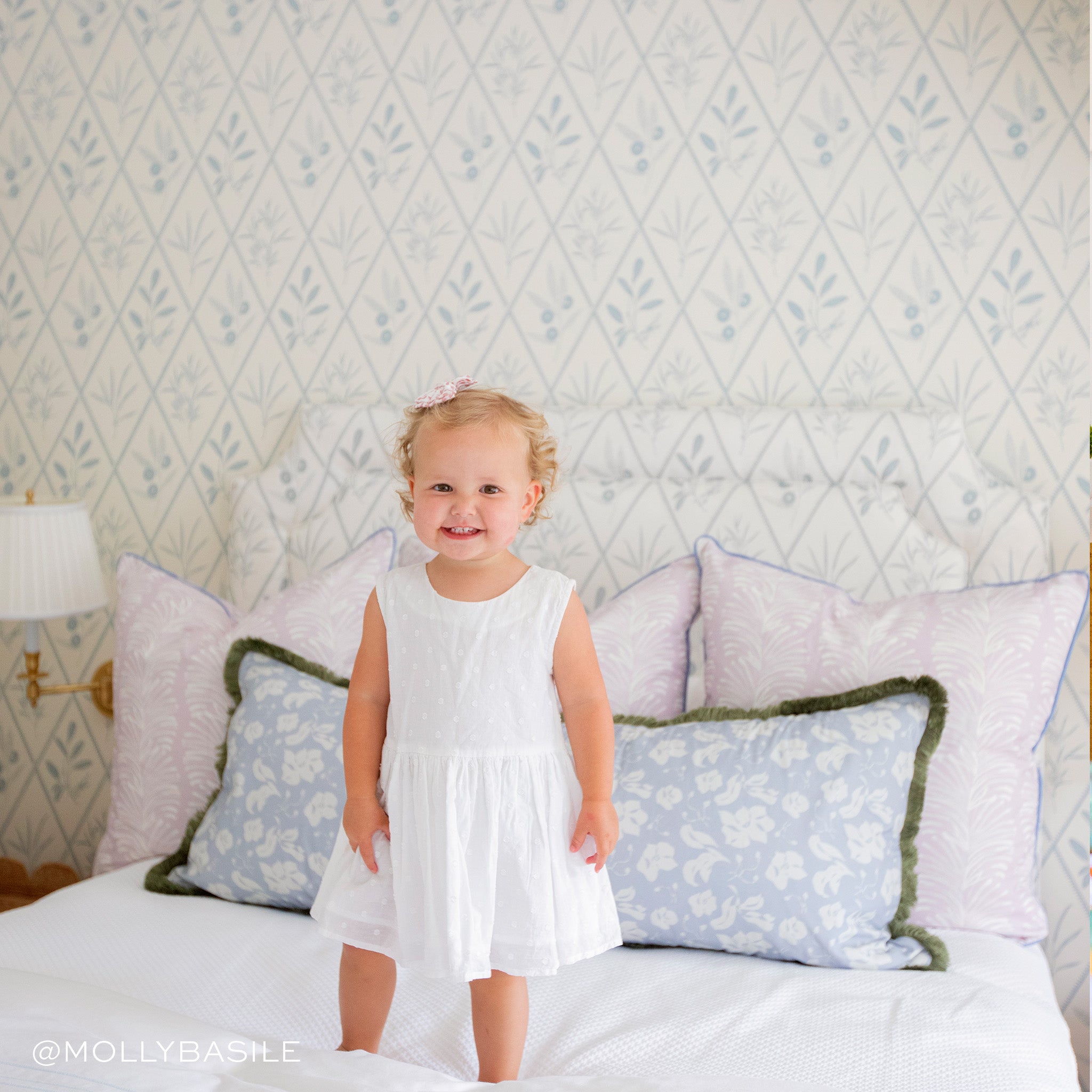 Bed styled with three Lavender Botanical Stripe Printed Pillows and one blue floral printed lumbar with sage fringe on white sheets with baby with a white dress and bow on the hair. Photo taken by Molly Basile