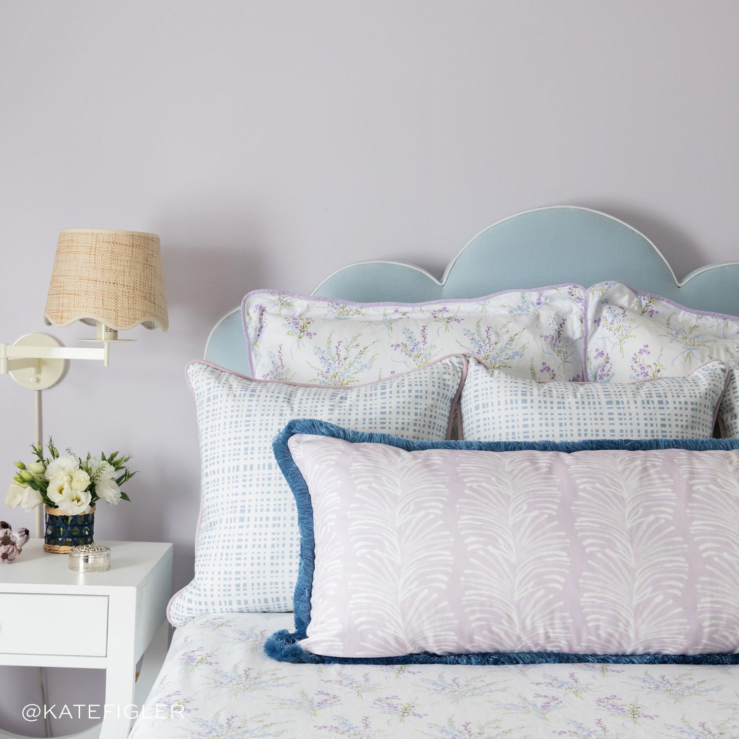 Bedroom styled with three blue gingham pillows and a Lavender Botanical Stripe Lumbar with blue fringe on bed next to white nightstand with lamp and flowers. Photo taken by Kate Figler