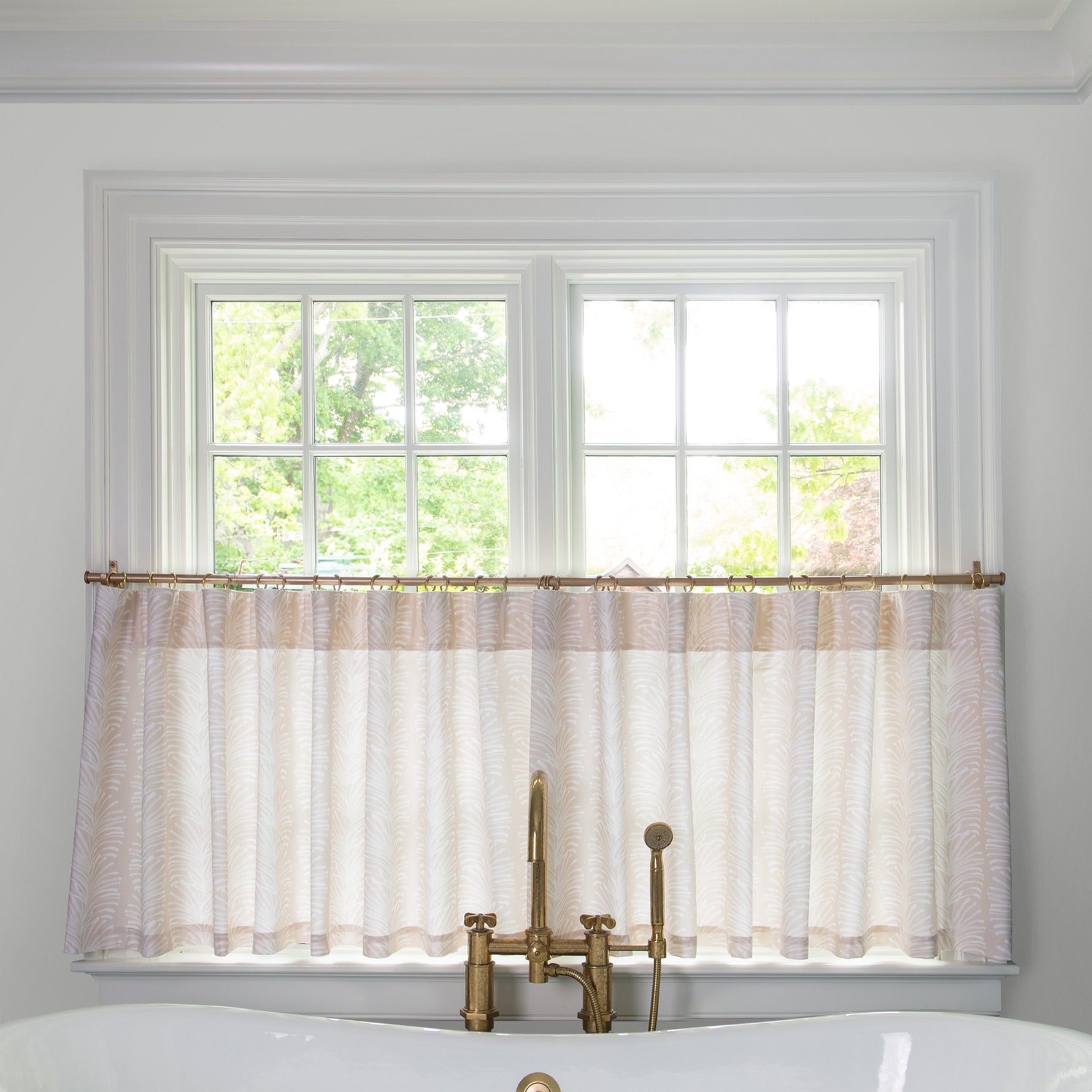 beige Botanical Stripe cafe curtain hung in front of an illuminated window in a bathroom with a white bathtub in front of the window with a gold faucet 