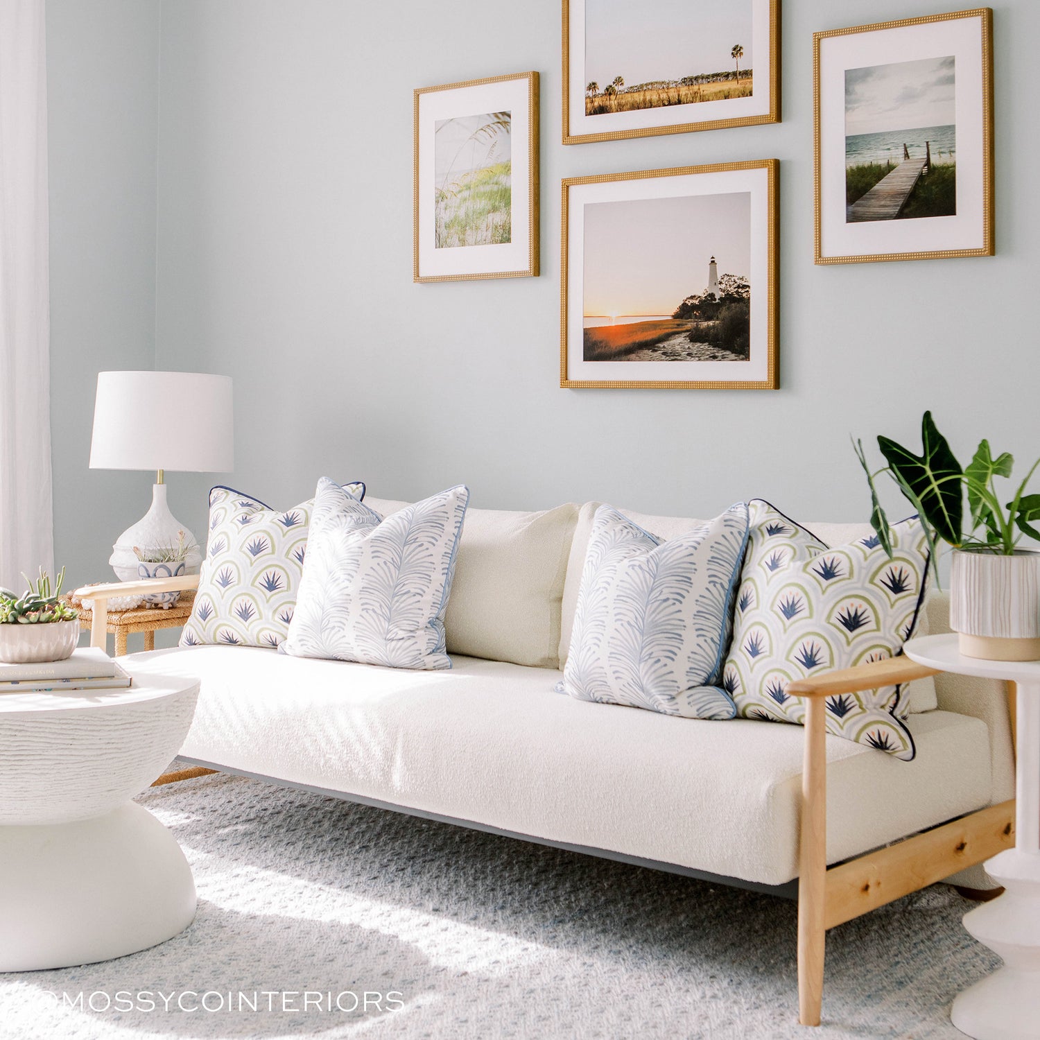 room with white couch with blue botanical stripe pillows and blue and green art deco palm pillows on the couch, photos of beach landscapes in brown wooden frames on the wall above the couch. a white circular coffee table with books and a plant on it 