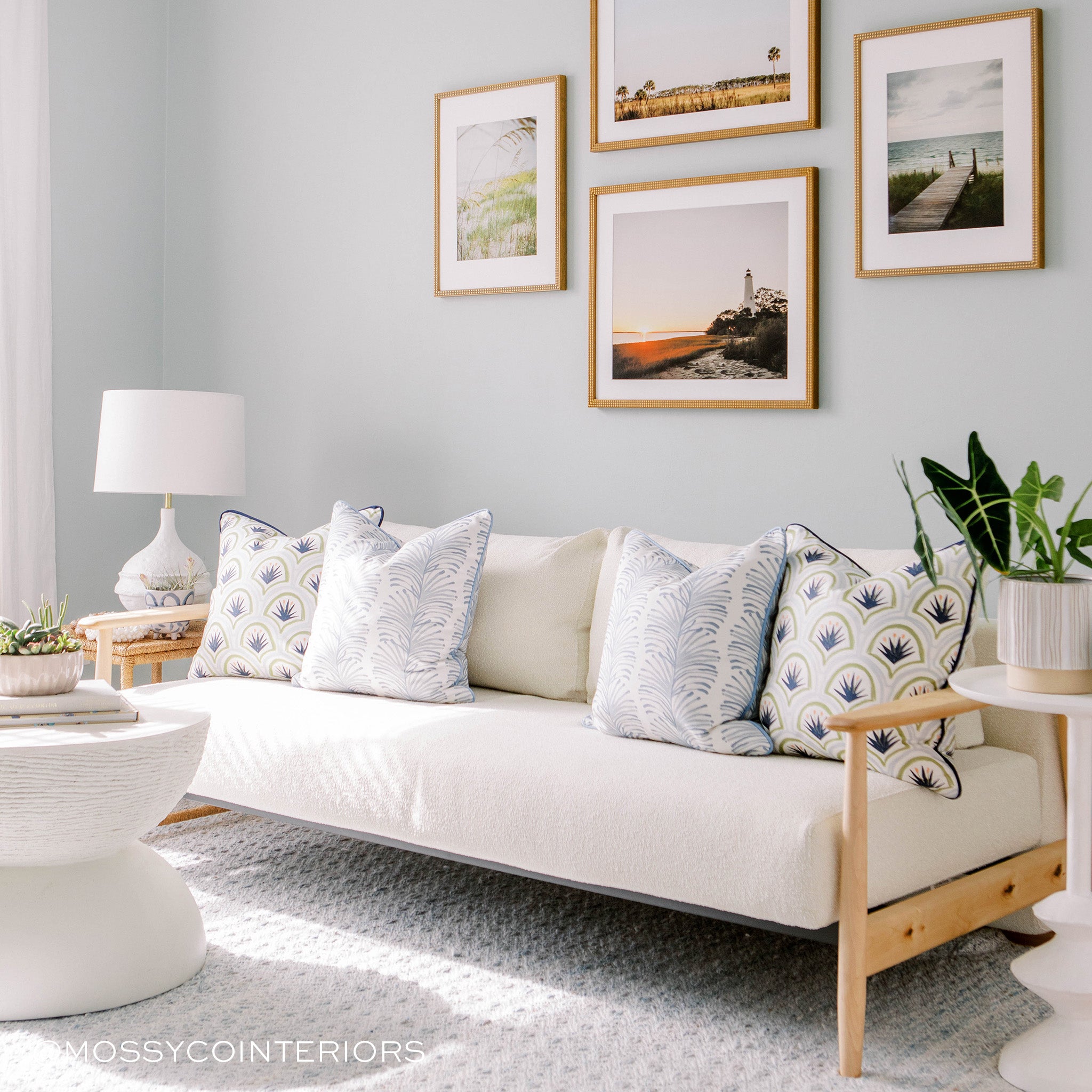 room with white couch with blue botanical stripe pillows and blue and green art deco palm pillows on the couch, photos of beach landscapes in brown wooden frames on the wall above the couch. a white circular coffee table with books and a plant on it 