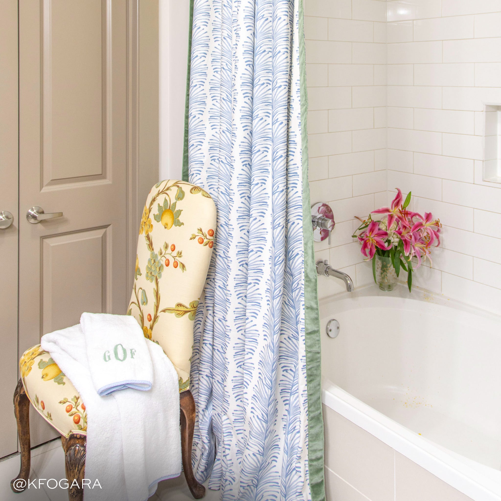 sky blue botanical stripe patterned shower curtain with a white tiled bathtub with flowers inside of the tub and a floral chair in front of the tub