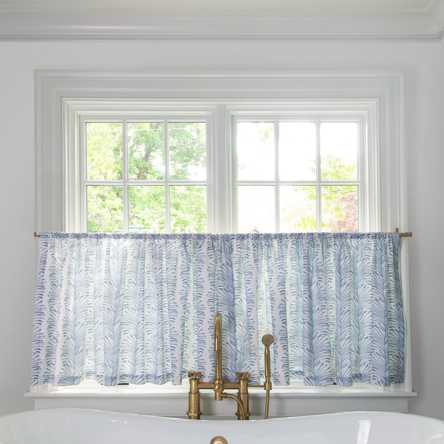 Sky Blue Botanical Stripe cafe curtain hung in front of an illuminated window in a bathroom with a white bathtub in front of the window with a gold faucet 