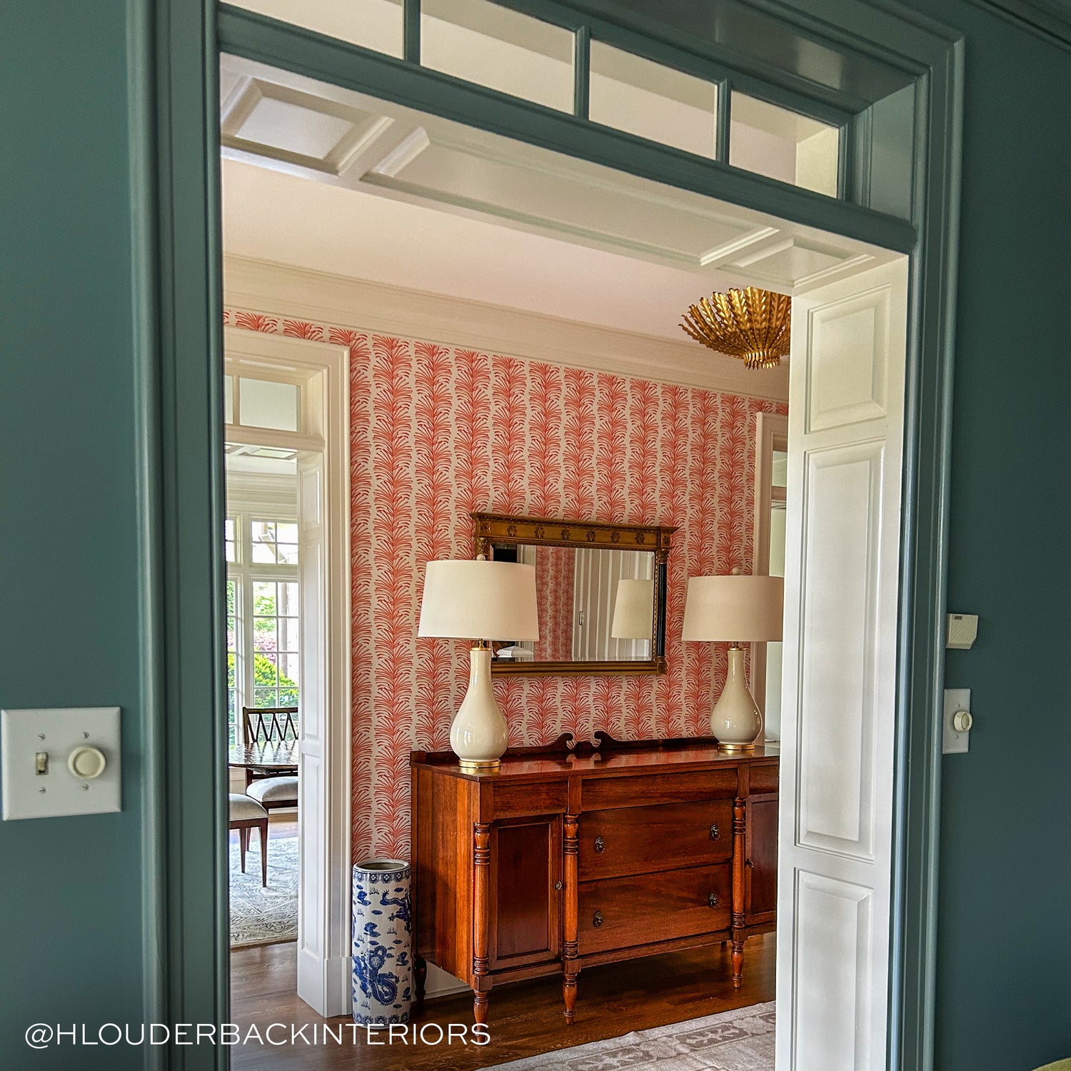 photo of an entry way with coral botanical stripe wall paper on the walls a gold light fixture and a dark wooden chest against a wall with two white lamps on it 