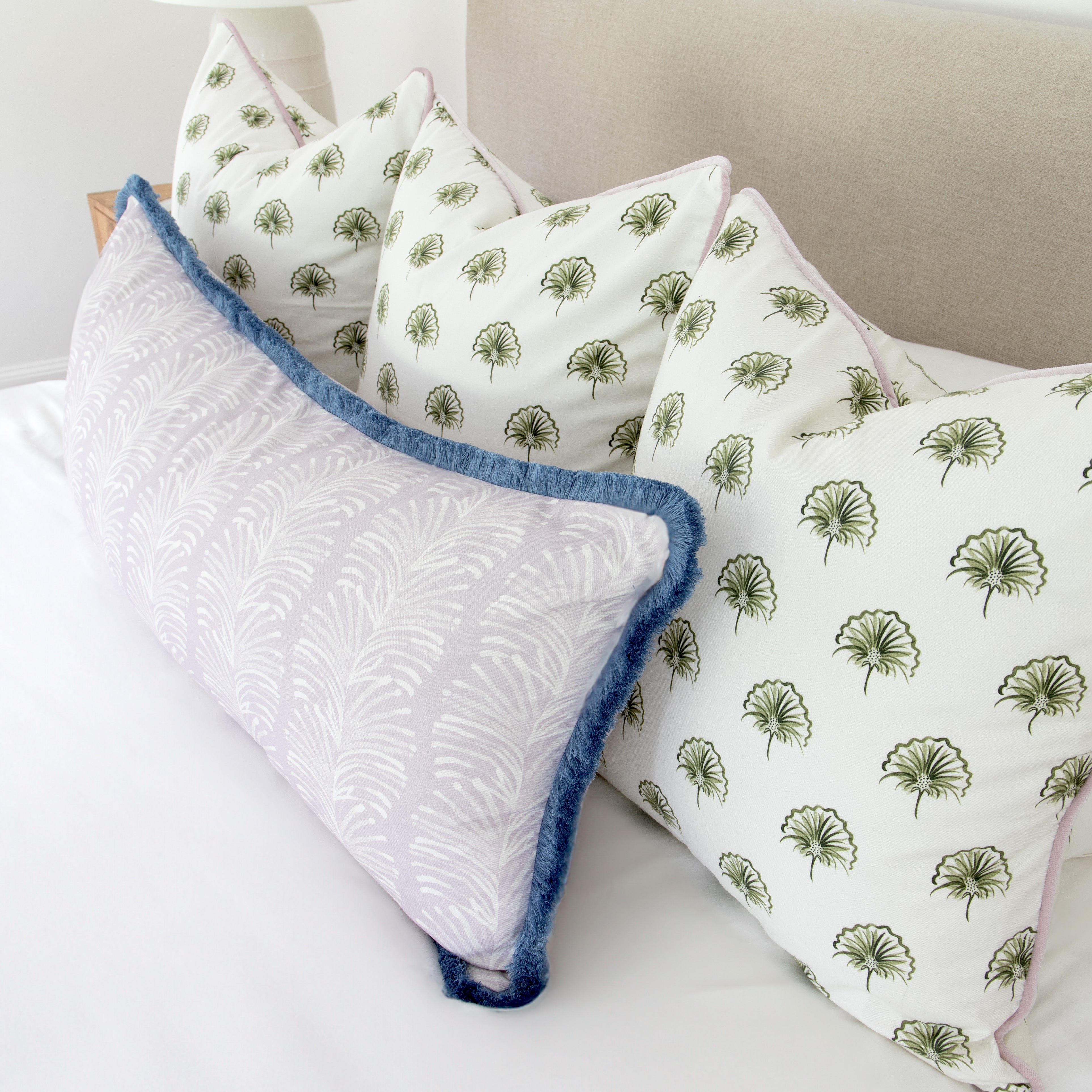 Bedroom close-up with three green floral printed pillows and one Lavender Botanical Stripe printed lumbar with blue fringe on top of white bed 