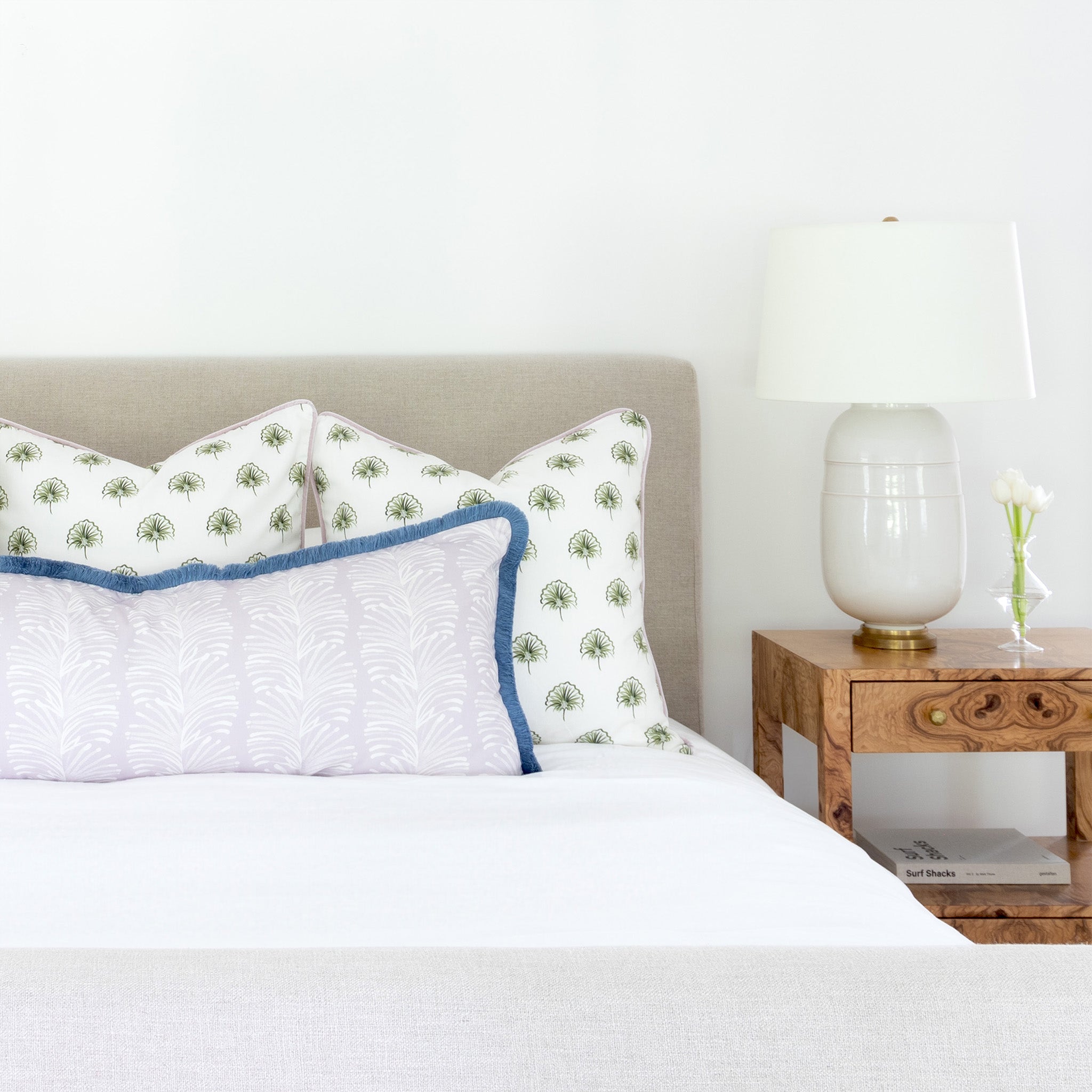 Bedroom close-up with two green floral printed pillows and one Lavender Botanical Stripe printed lumbar with blue fringe on top of white bed next to brown table with white lamp on top