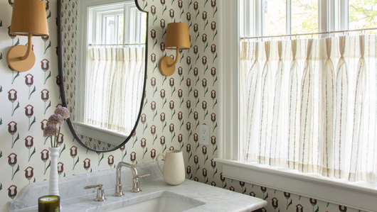 neutral background wallpaper with pink and maroon wallpaper in a bathroom with sheer white cafe curtains embroidered with white stripes hung in front of an illuminated window 