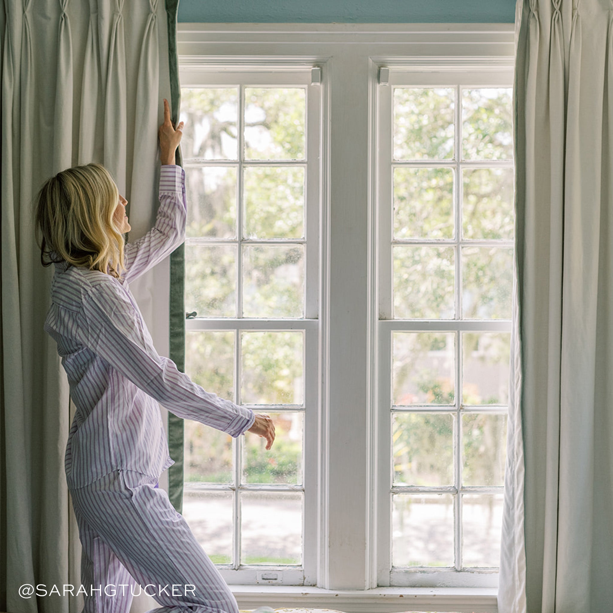 Illuminated window styled with Natural White Linen Curtains with Fern Green Velvet Band being held by blonde woman wearing stripped pajamas. Photo taken by Sarah G Tucker