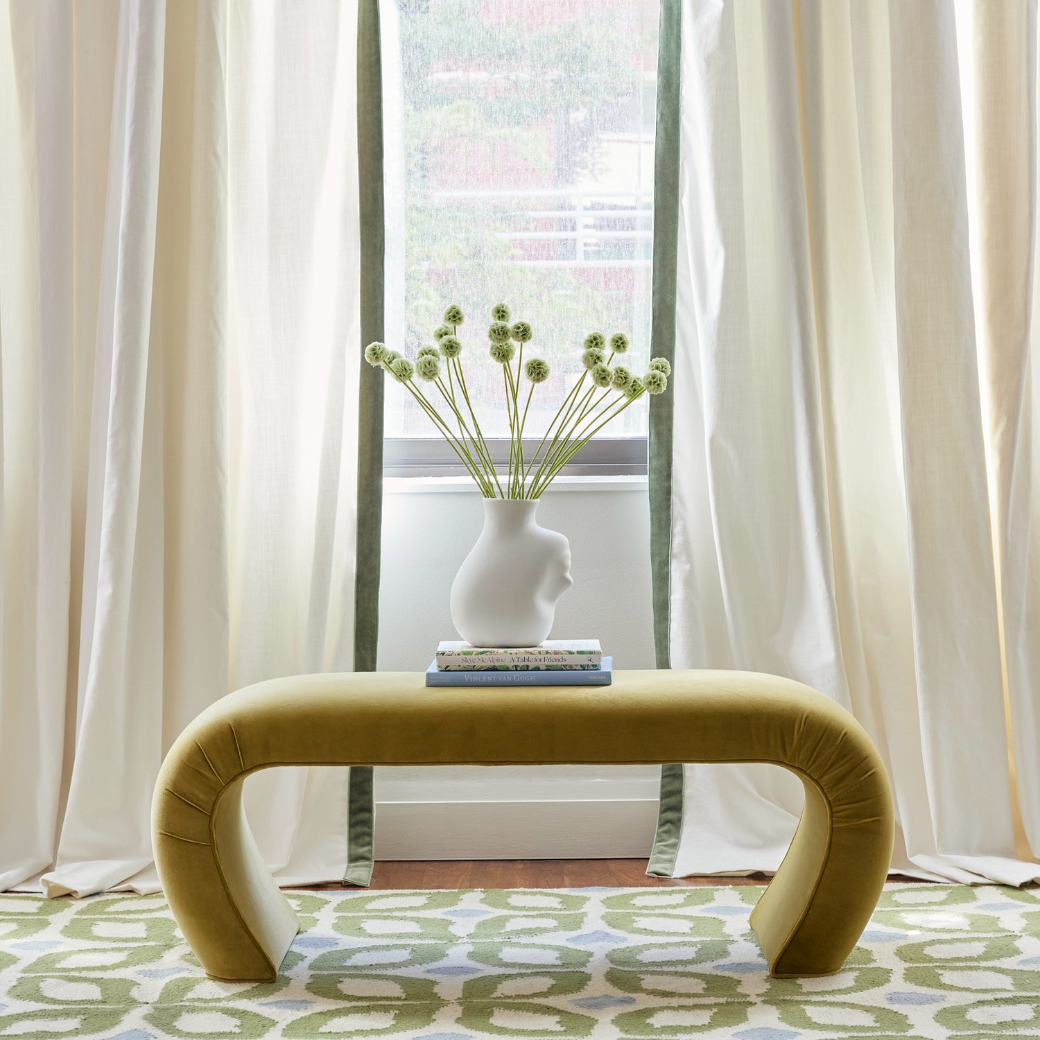 Close-up of illuminated window styled with Natural White Curtains with green velvet band by green plants in white vase on top of stacked books on mustard yellow velvet bench over Moss Green Geometric Printed Rug 