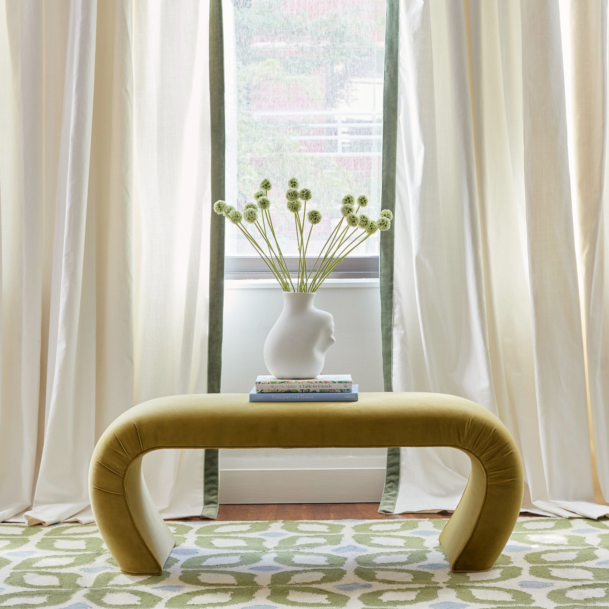 Close-up of illuminated window styled with White Cotton Curtains with green velvet band by green plants in white vase on top of stacked books on mustard yellow velvet bench over Moss Green Geometric Printed Rug 