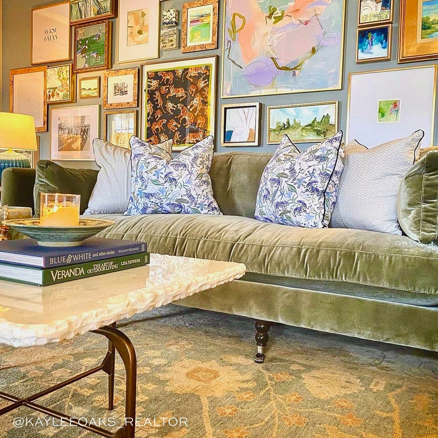 Living room full with artwork on wall styled with two Green Tiger Printed Pillows facing each other and two Moss Green Geometric Printed Pillows on Green Velvet Couch next to cream table with candle on plate on two stacked books. Photo taken by Kaylee Oaks