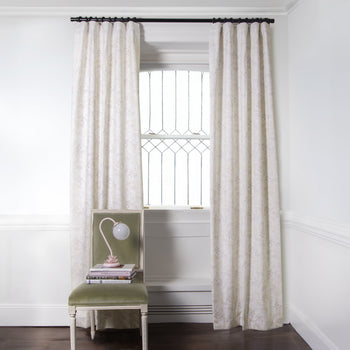 Beige Chinoiserie Tiger Printed Curtains on metal rod in front of an illuminated window with Green Velvet chair with floral pink lamp and stacked books on top