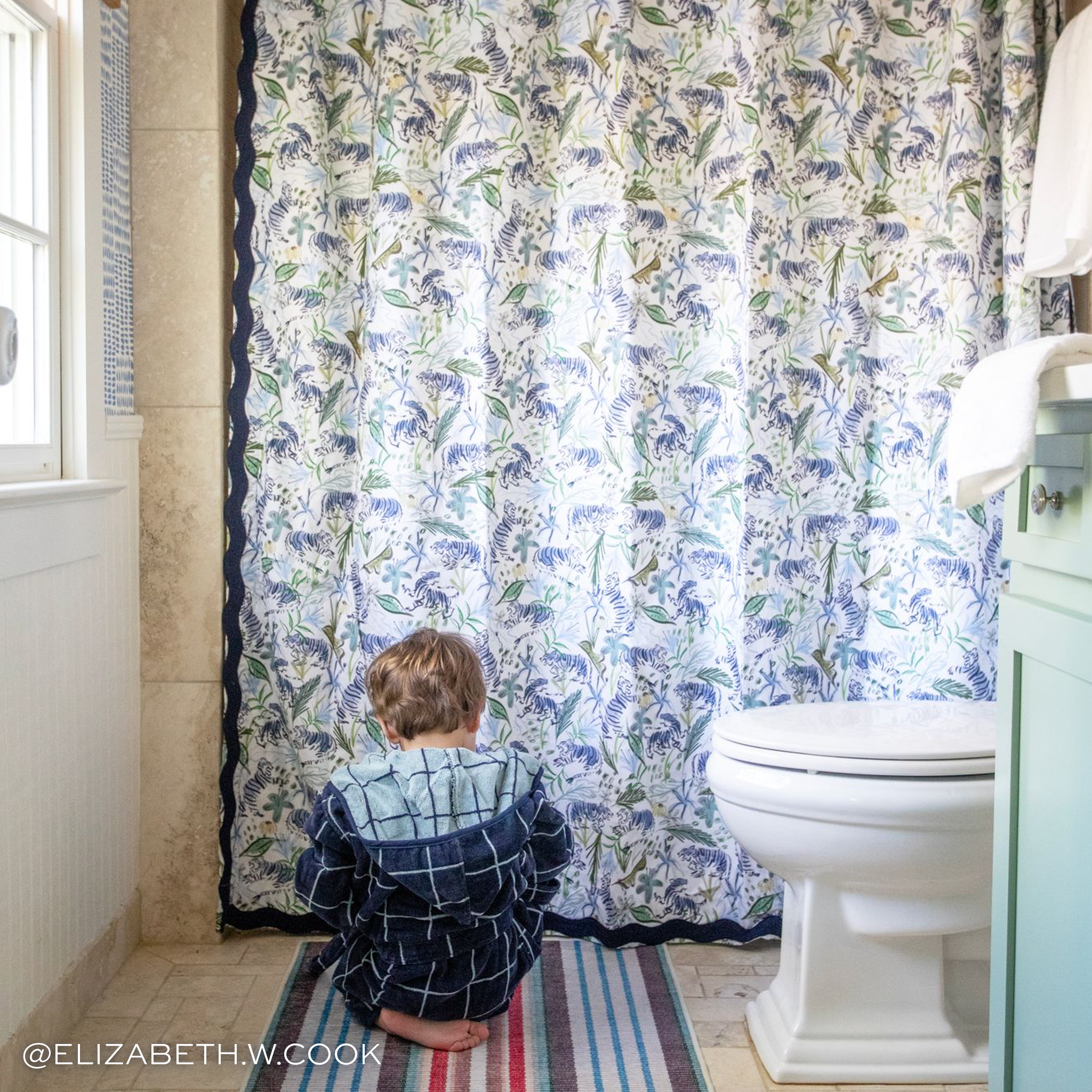 Green Tiger shower curtain hanging on rod in front of white tub in bathroom with little boy in a navy bathrobe sitting in front of the bath tub