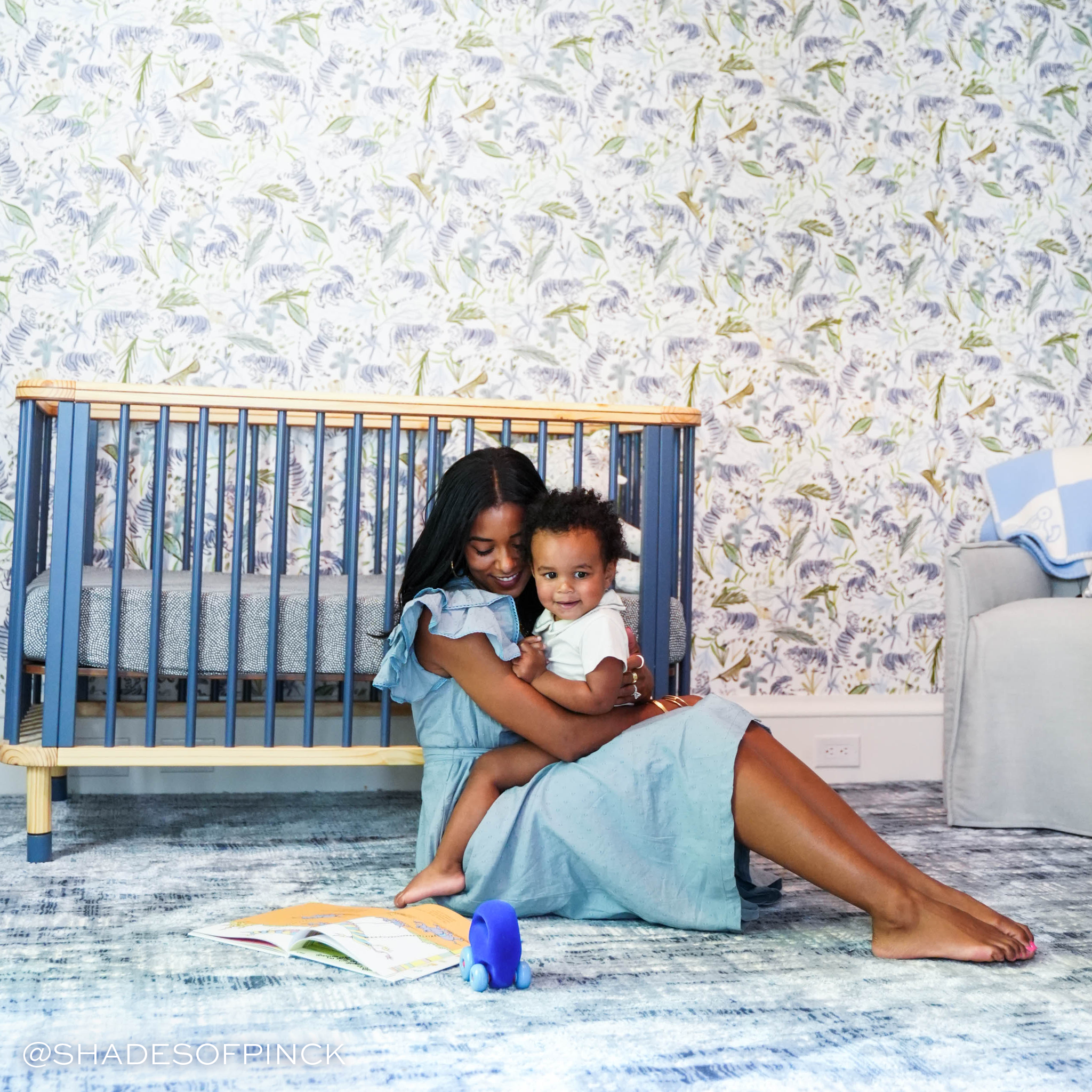 Nursery with mom and baby in front of a blue crib on a blue carper and Green Tiger Printed Wallpaper on the wall