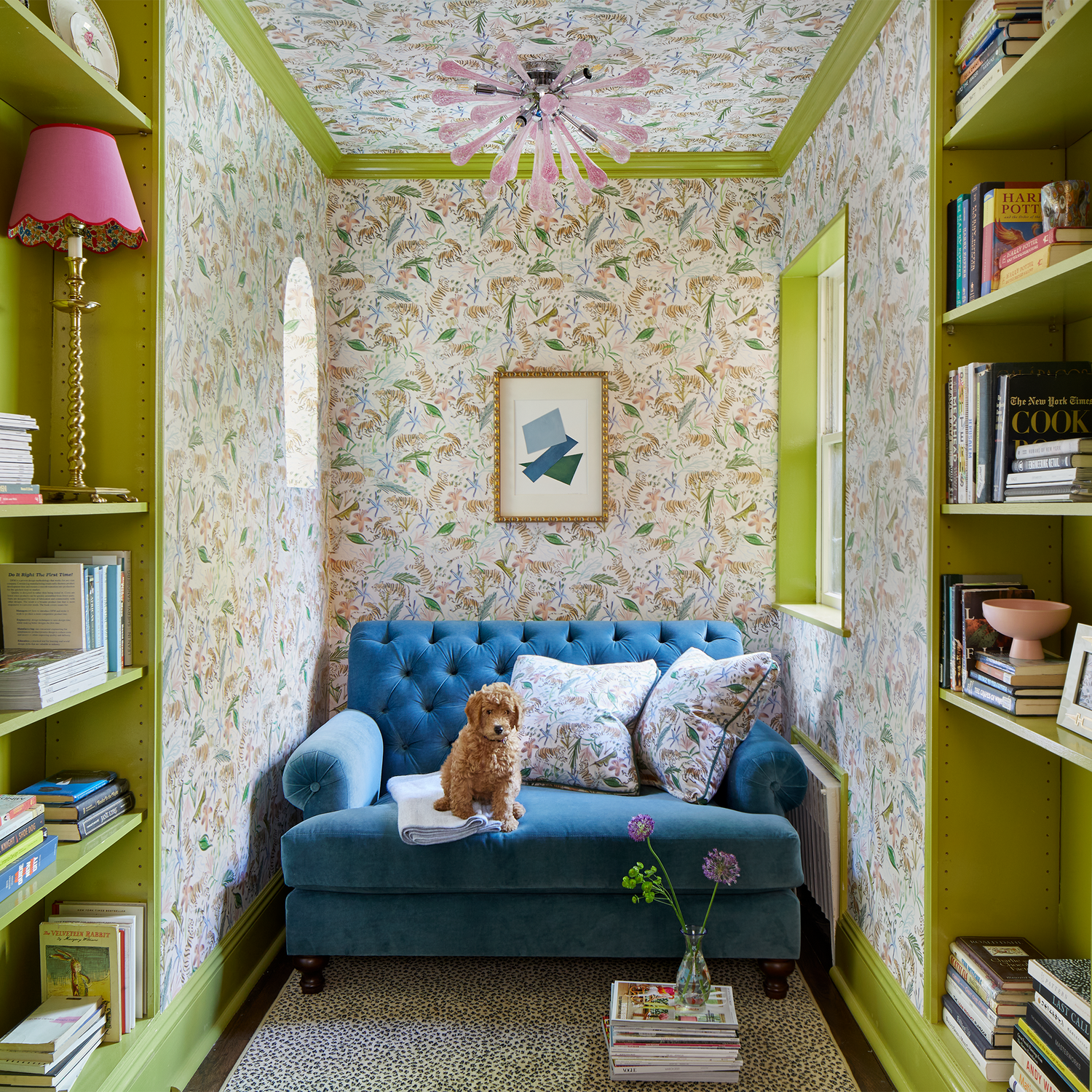 Library corner with books on shelves styled with Pink Chinoiserie Tiger Printed Wallpaper and blue couch with brown dog and Pink Chinoiserie Tiger Printed Pillows on top