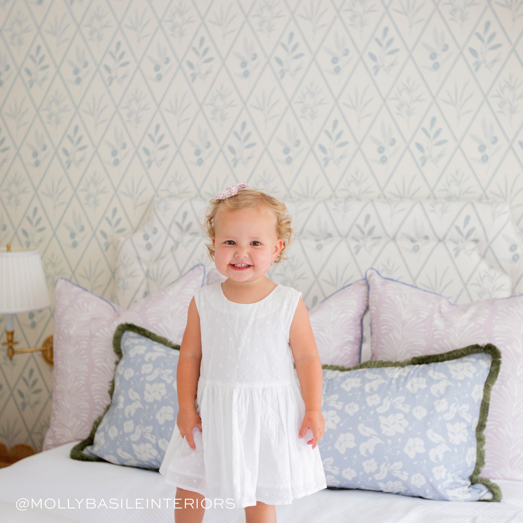 Bed styled with three Lavender Botanical Stripe Printed Pillows and one blue floral printed lumbar with sage fringe on white sheets with baby with a white dress and bow on the hair. Photo taken by Molly Basile