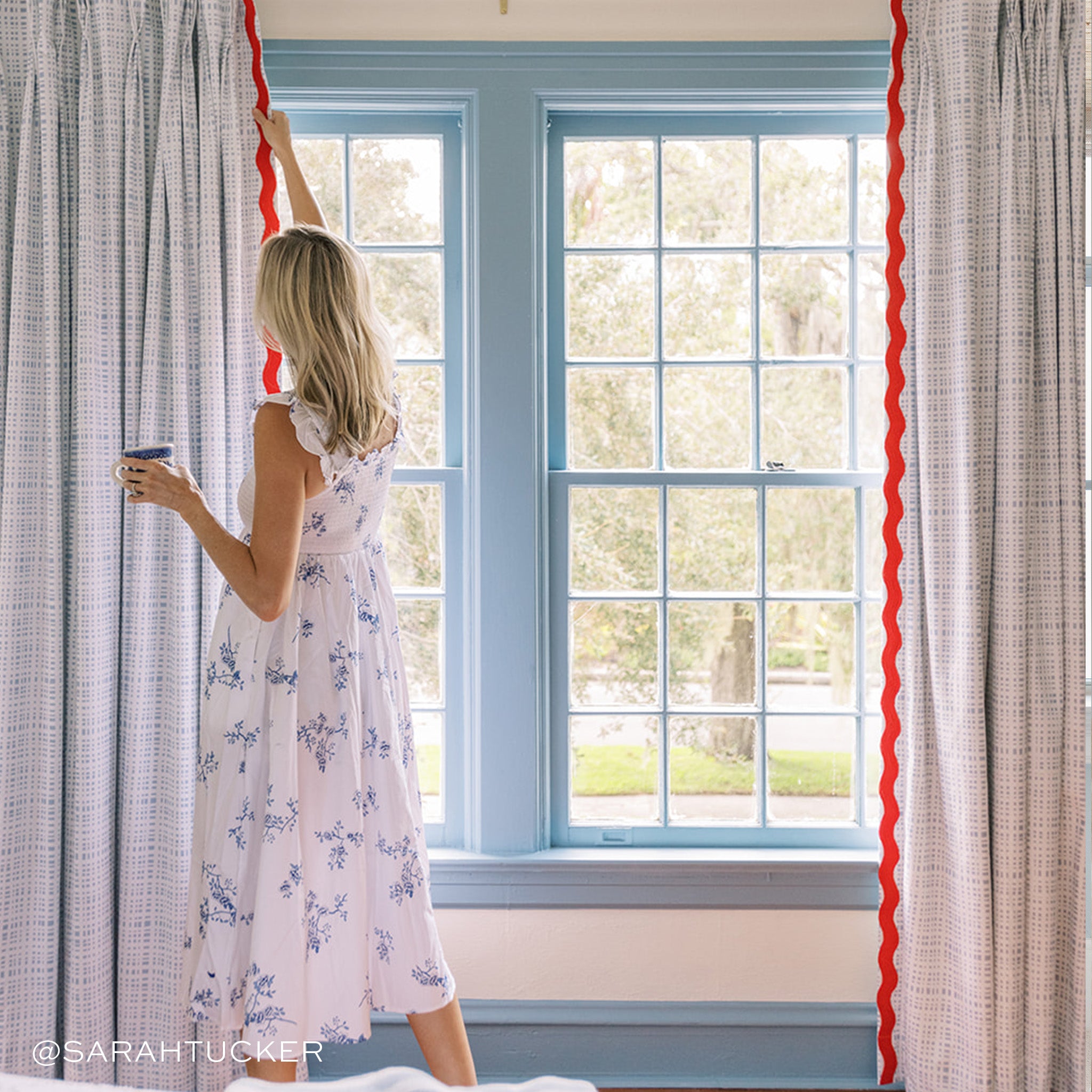 Illuminated window close-up styled with Sky Blue Gingham Printed Curtains with red band being held by blonde woman wearing a white and blue floral dress. Photo taken by Sarah Tucker