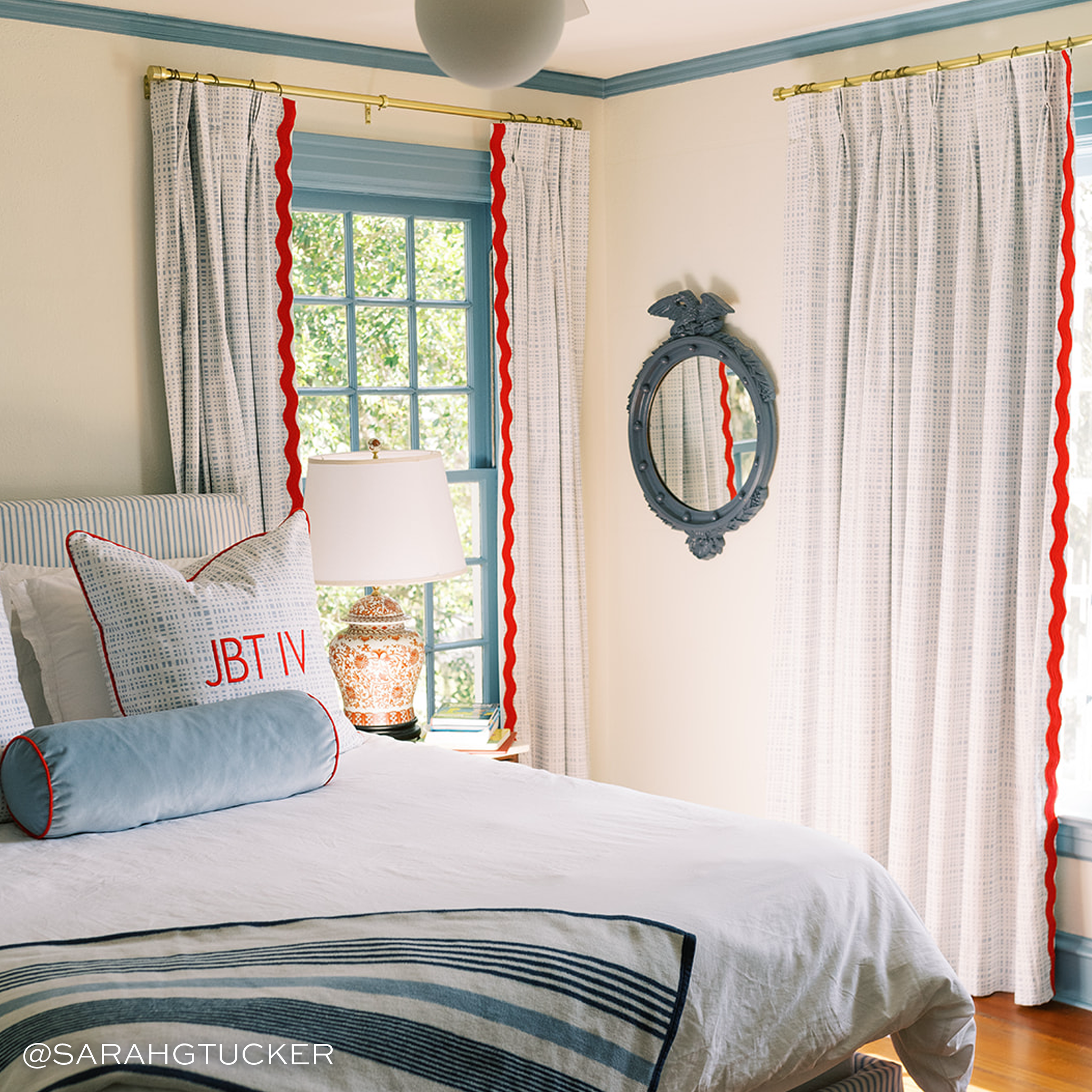 light blue plaid pillows with red trim and a solid light blue velvet pillow in front with red trim on a white bed in front of three blue plaid curtains with red zig zag trim
