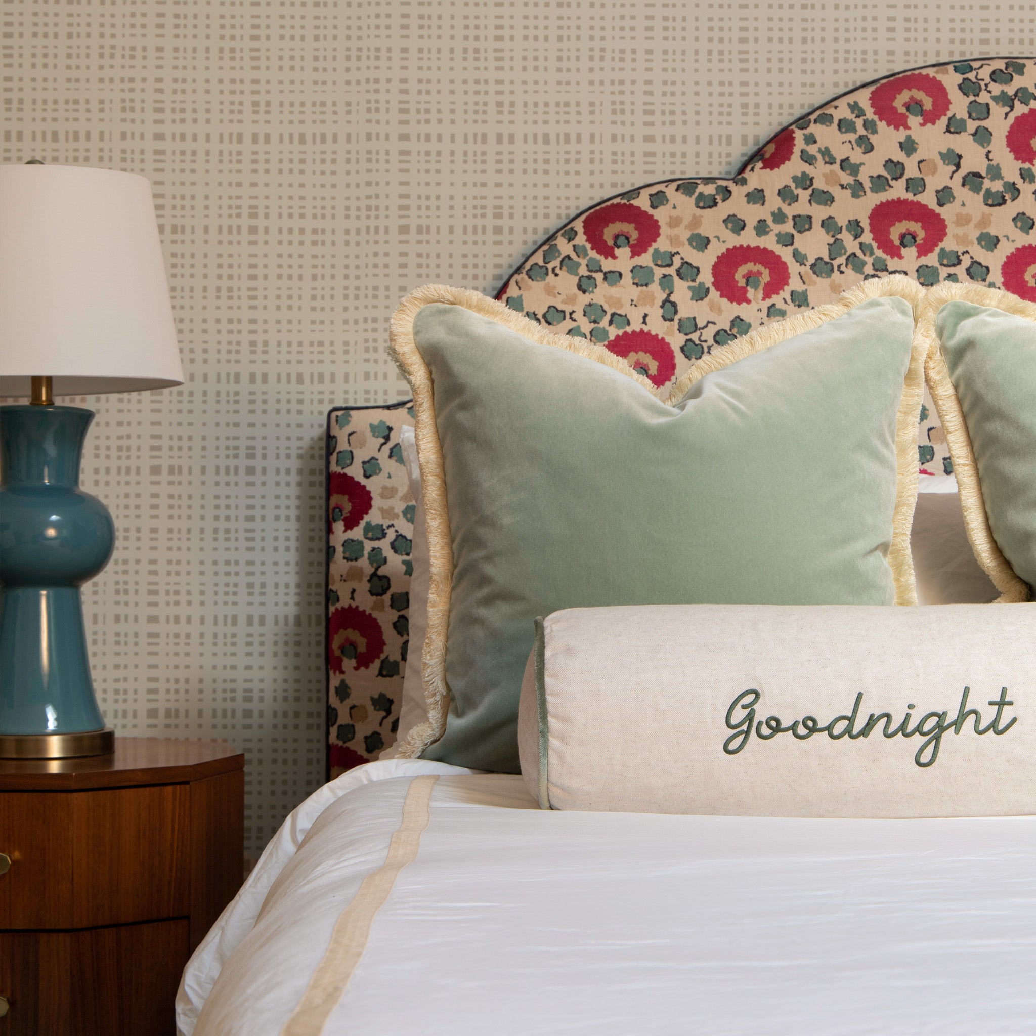 Close-up of white bed styled with two Blue Green Velvet Pillows and one Light Brown Monogrammed Linen Lumbar next to wooden nightstand with blue and white lamp on top in front of beige gingham printed wallpaper