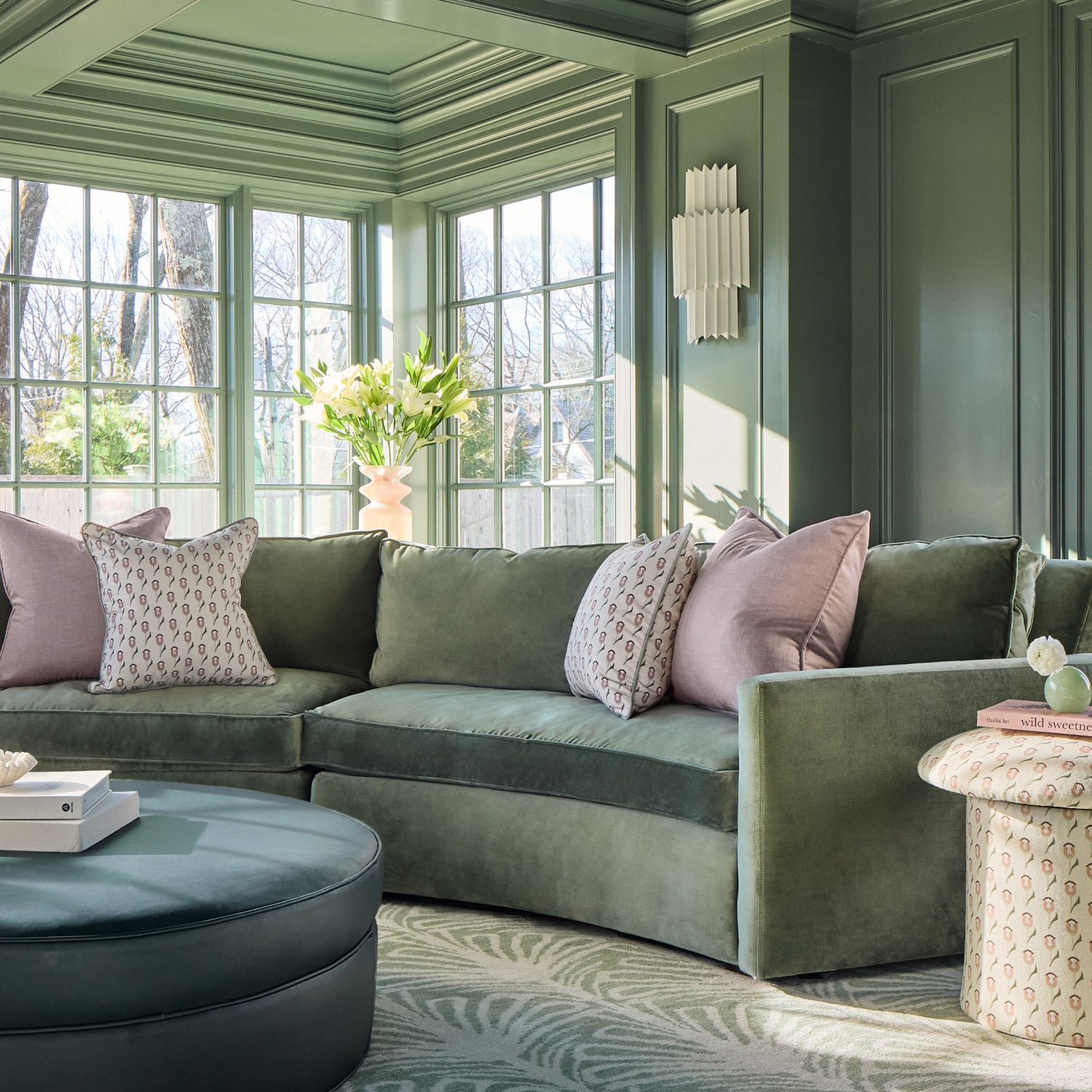 abstract floral pink and green pillows on a green couch with a blue ottoman with books stacked on it and green painted walls