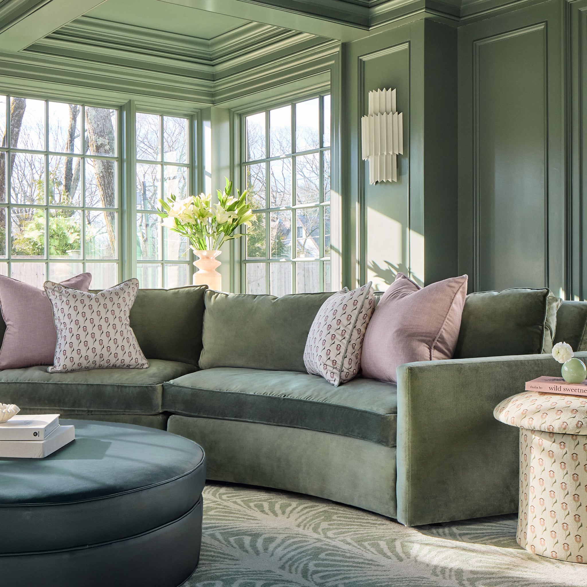 soft pink pillows on a green couch with floral pillows and a blue ottoman stacked with books in a room with green walls